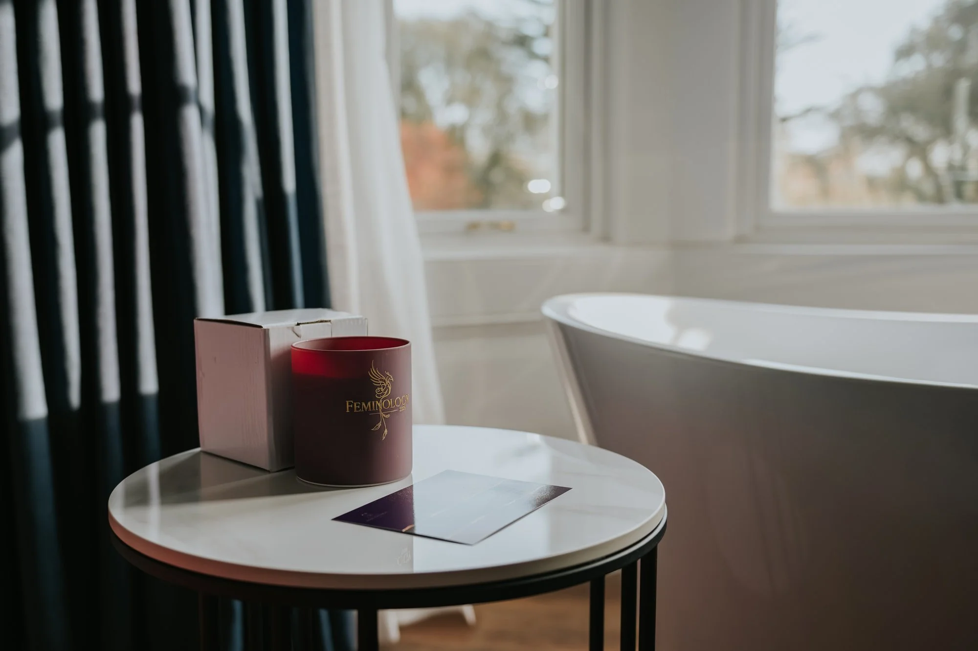 A wide shot of a purple candle jar sitting on a table, the word 'Feminiology' are written in gold across it. Behind it is a big freestanding bath in front of a big bay window.