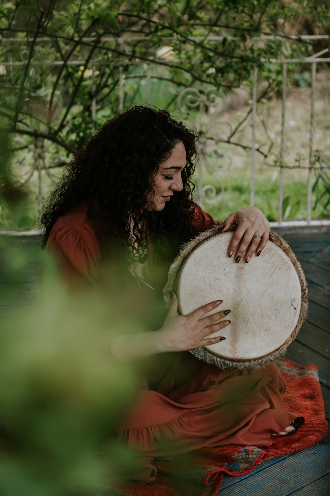 El Jedras, woman dressed in a flowy orange dress, sitting on a pergola. She is surrounded by greenery and is lovingly looking at her drum.