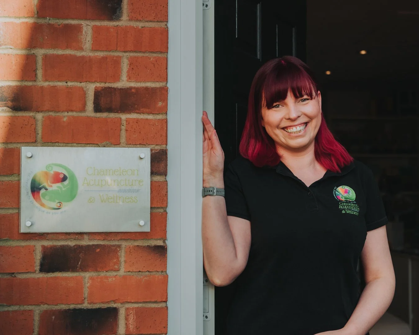 Vikki Chameleon Acupuncture. Woman with bright red hair standing in the door way next to her business sign. She is smiling brightly at the camera.