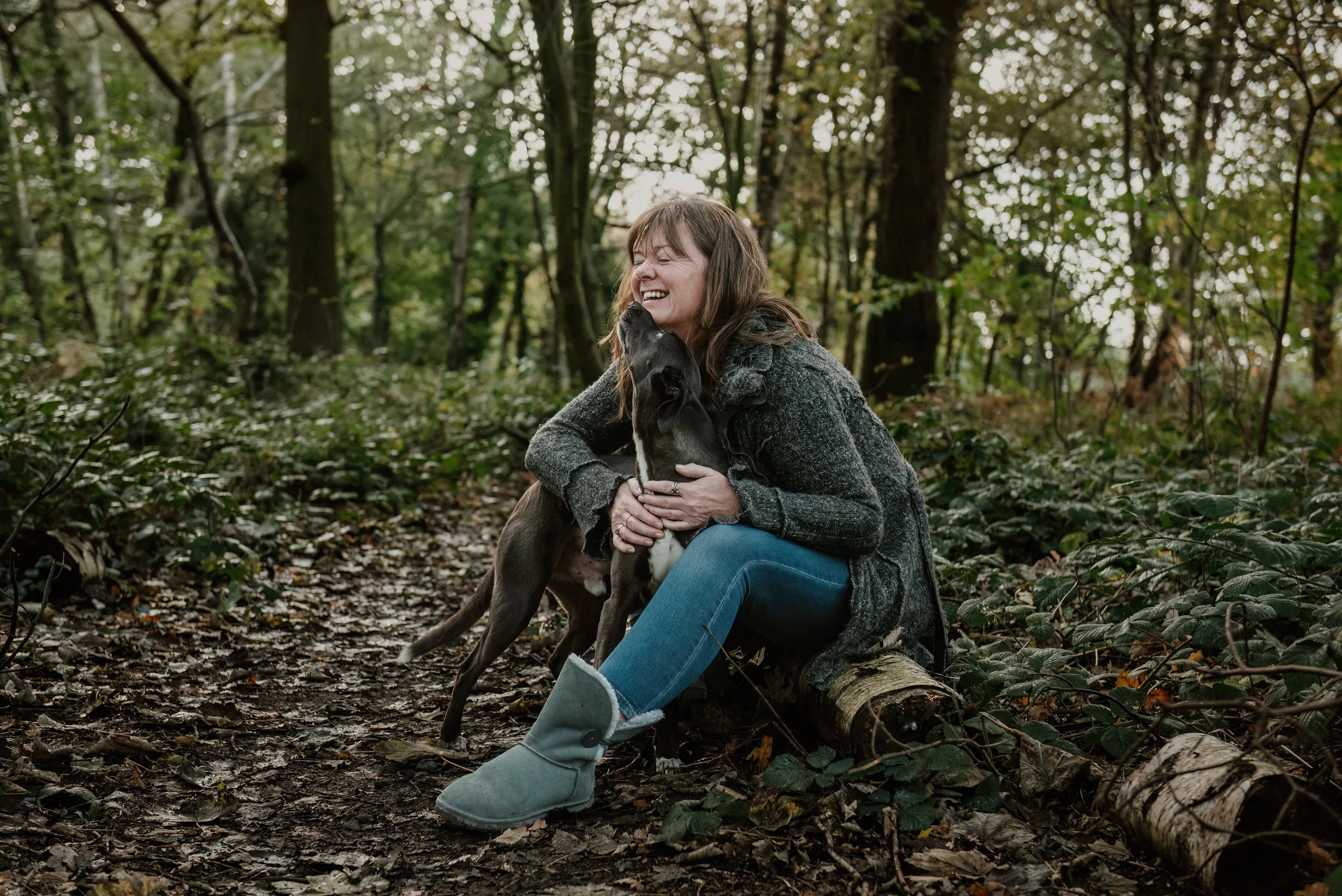 Paula, Tiny Rituals. She is sitting on a log in the woods with her beautiful dog Buddy who is looking up and touching her face with his nose. She has her eyes closed and is smiling.