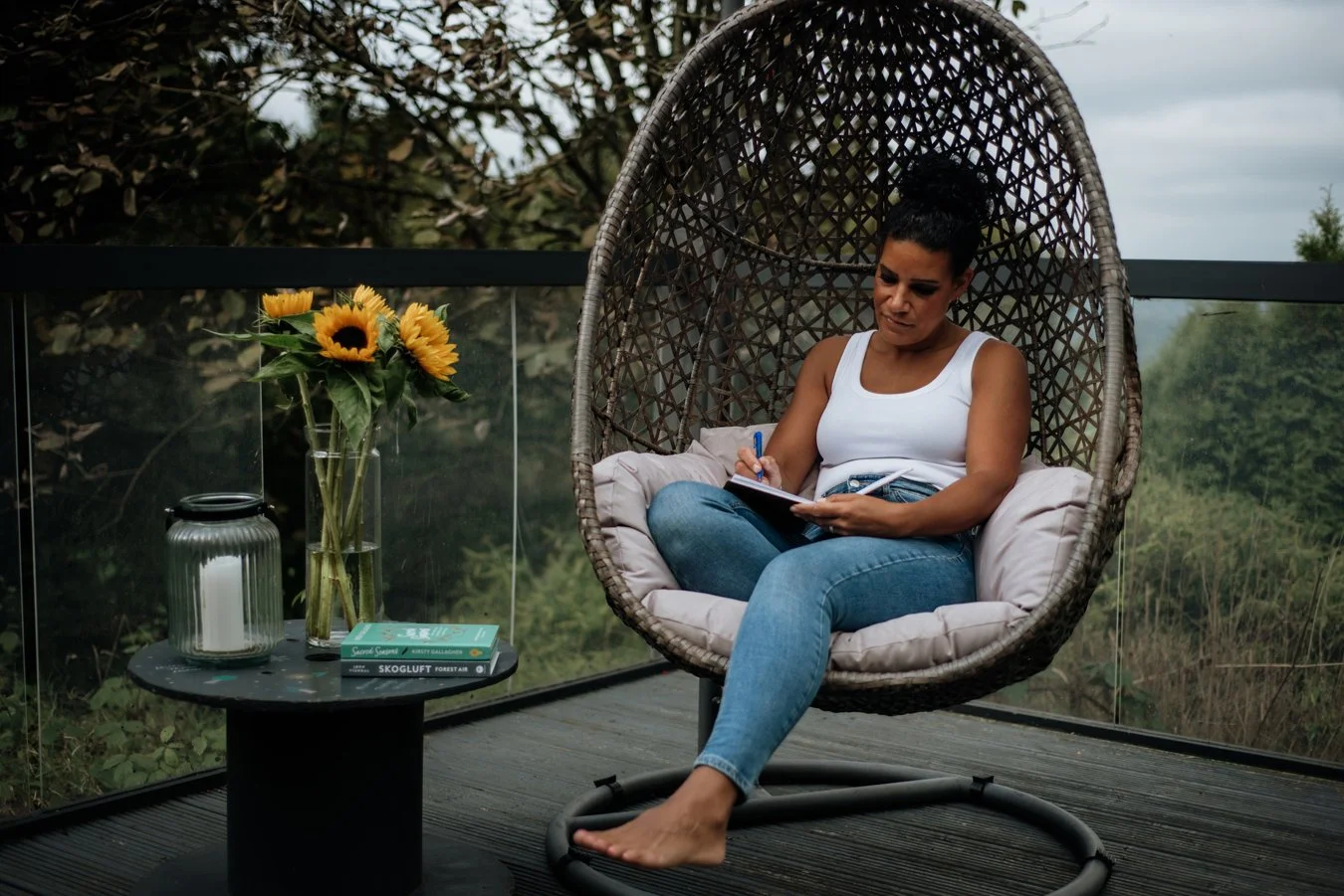 Woman sitting in a suspended chair outdoors while writing in her journal. There are sunflowers beside her.