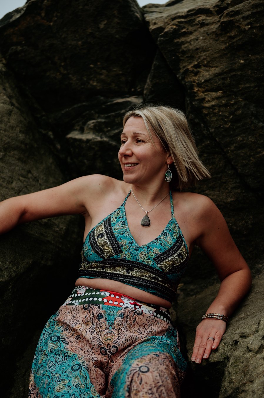 Kelly Bisson, Kundalini with Kelly. She is dressed in a beautiful patterned two piece and leaning against some dark rocks.