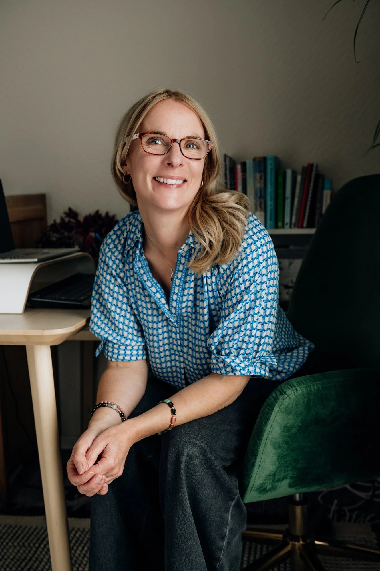 Sophie Longden Therapy. Sophie is dressed in blue and sitting in her office. She has her hands together and is leaning forwards smiling while looking up.