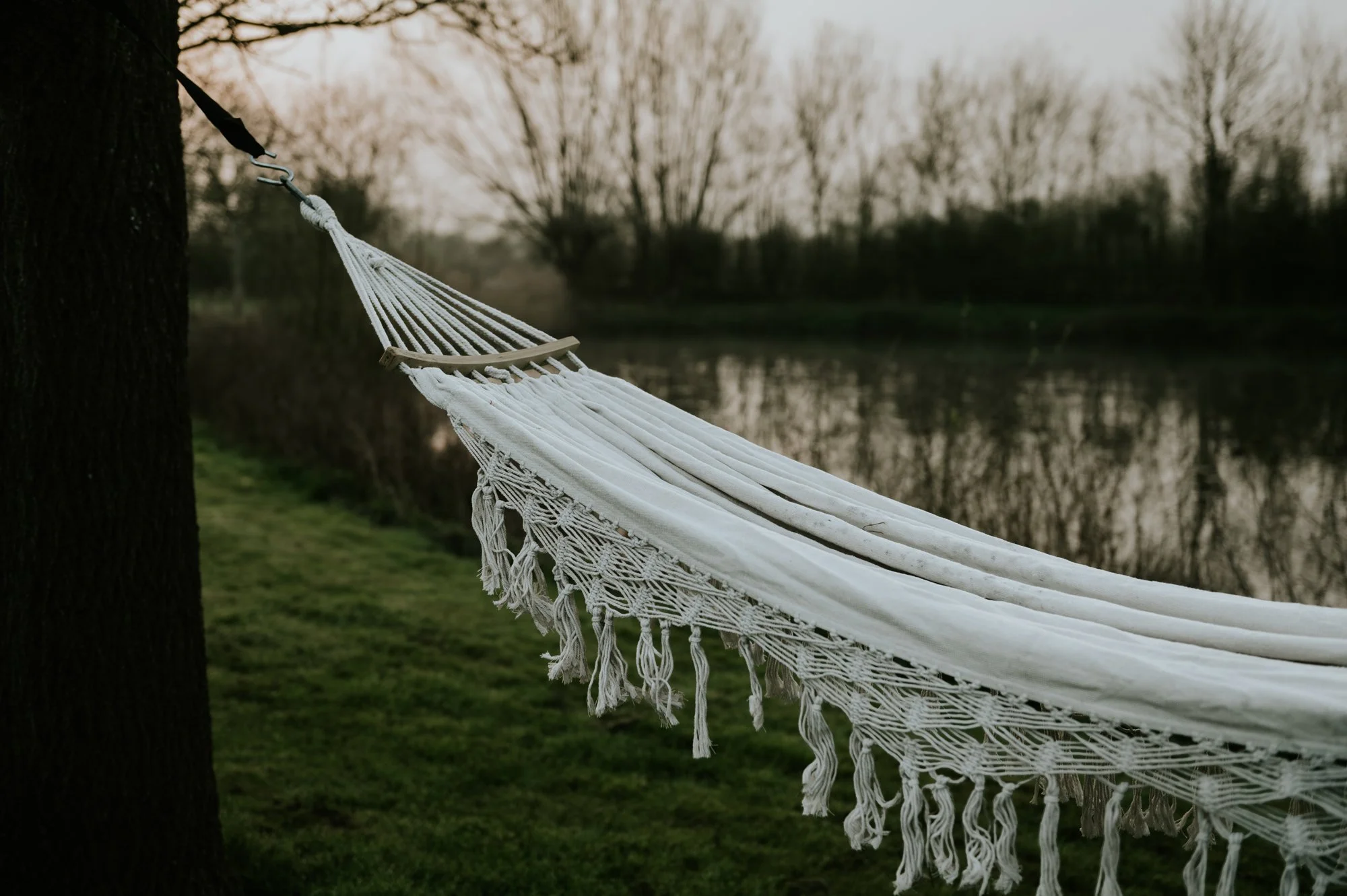 A white canvas hammock with tassels down the side of it, suspended on a tree, you can see a lake in the distance behind it.