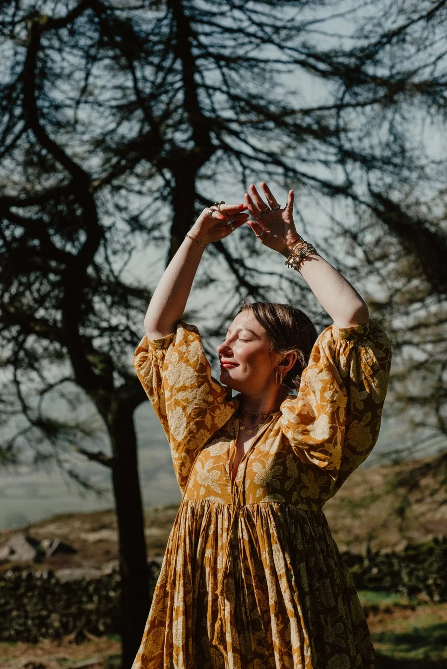 Molly Gould, Effable. She is reaching up into the air, there is a tree above her creating beautiful dappled light.