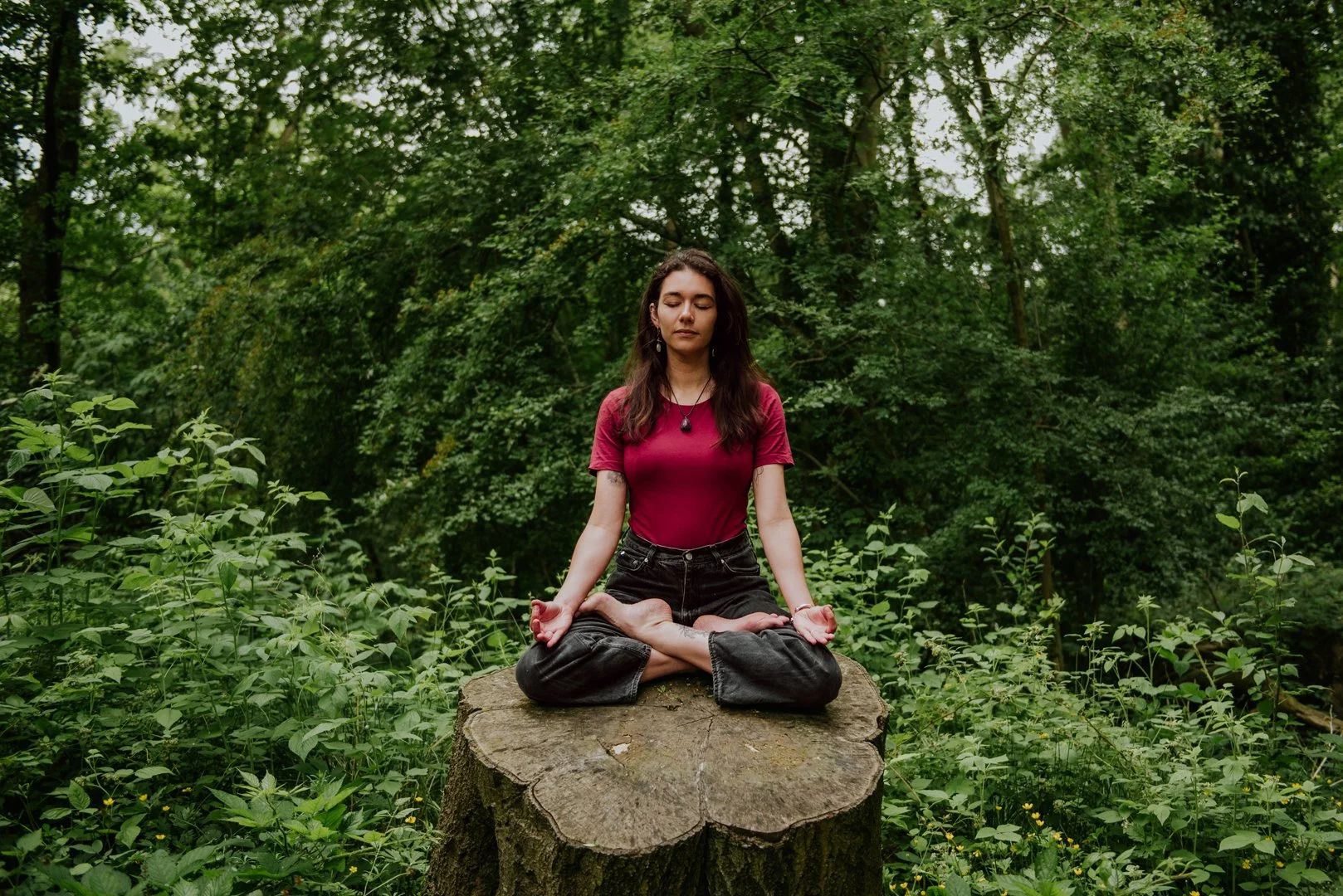 Ellie Belfield, therapist. She is sitting on top a cut off tree truck with her legs crossed in a yoga pose. Her eyes are shut in meditation.