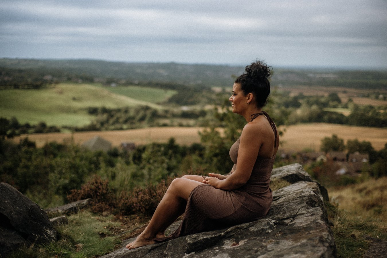 Nina, Nurtured Through Nature. Nina is sitting on rocks looking out over the landscape of hills and fields.