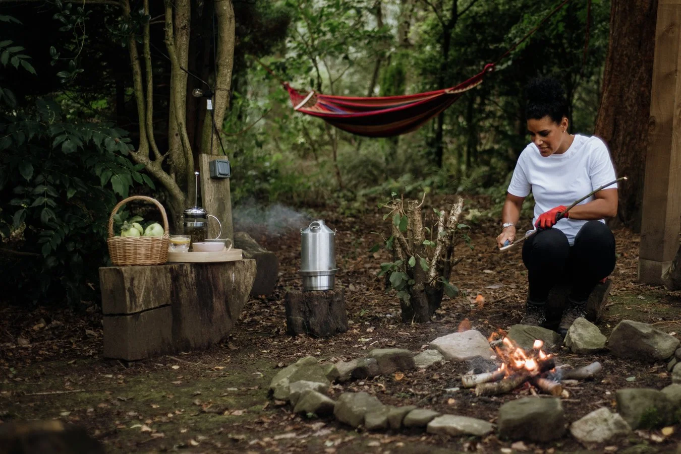Nina, Nurtured Through Nature. Nina is sitting by a fire and whittling a wooden stick. There is a hammock in the background and water brewing beside her.