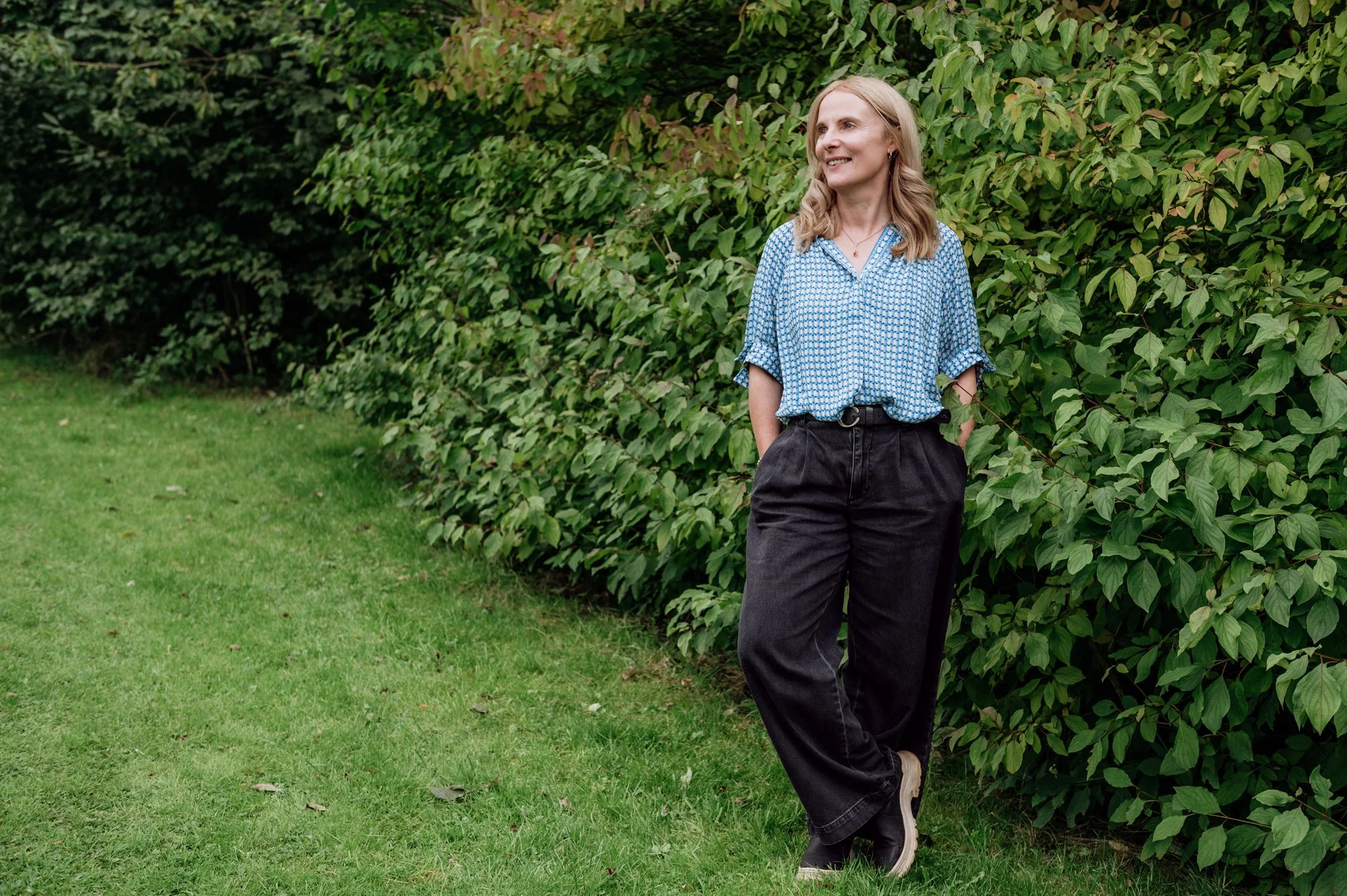 Sophie Longden Therapy. Sophie is standing casually in front of beautiful green bushes. She is looking to the side.