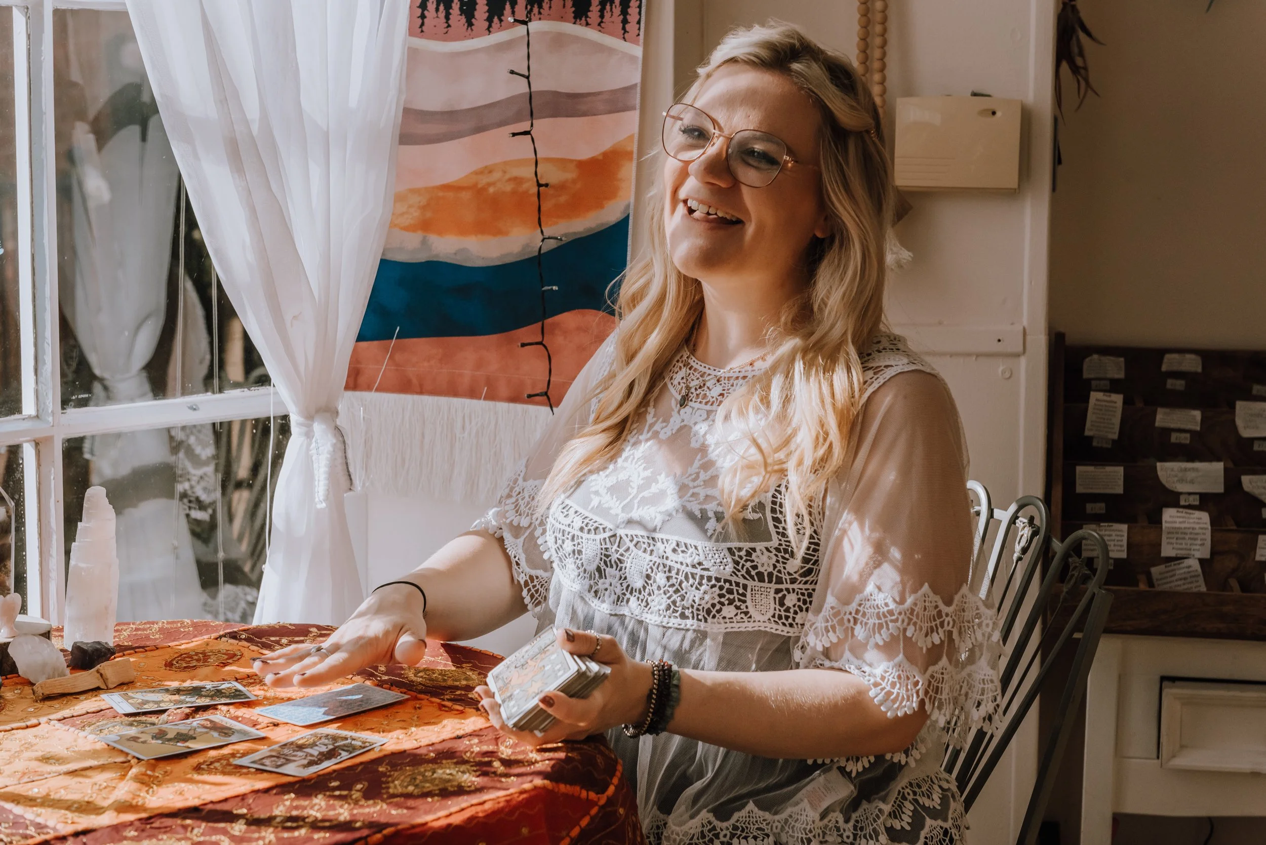 Camilla. Soul Reiki. She is dealing out Tarot cards while sitting at a table by a window. She looks happy.