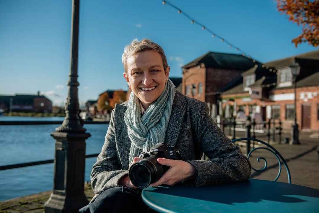 Dawn from DLT Visions is sitting at a table in the harbour. She is holding her camera and smiling.