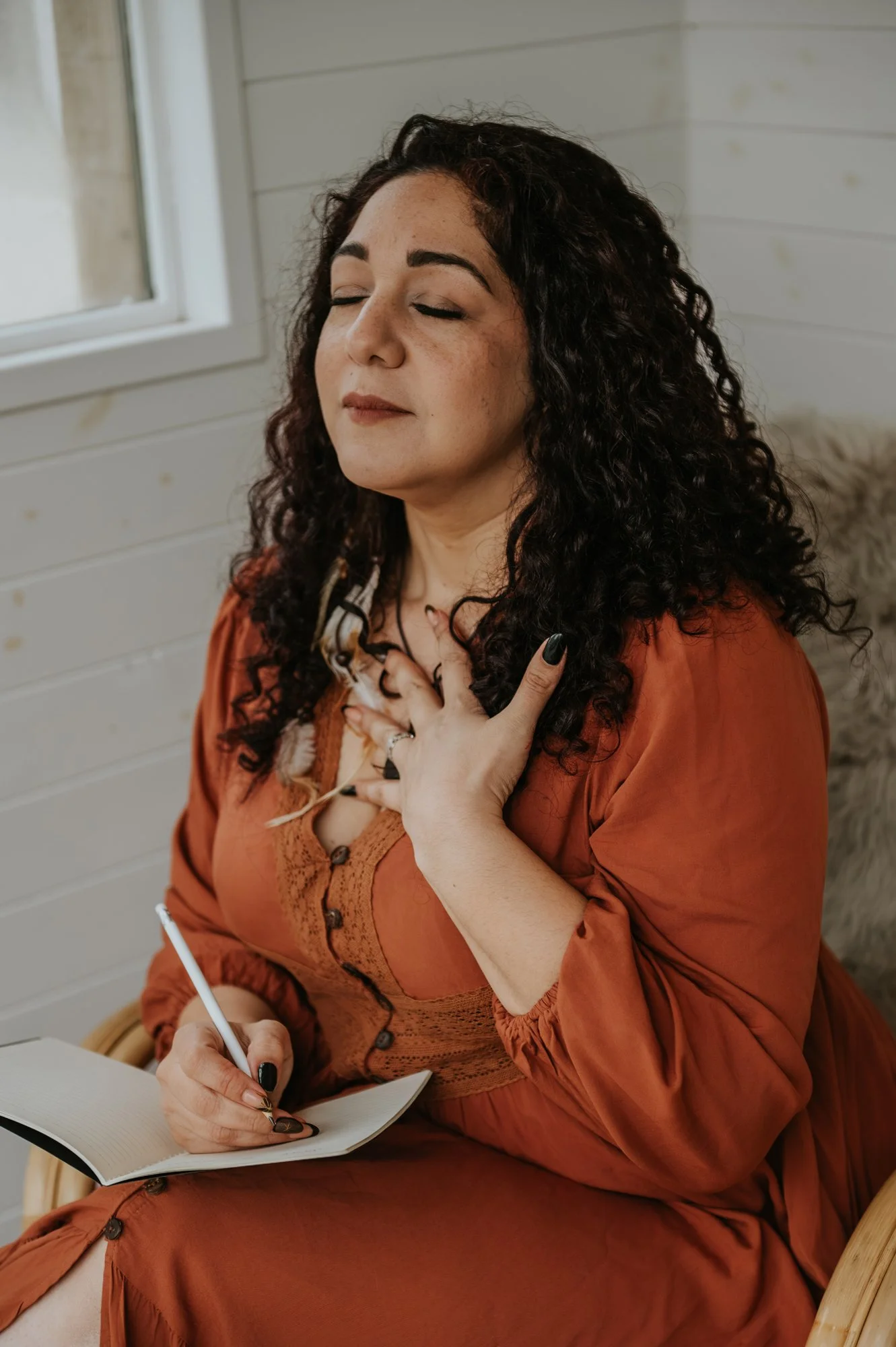 El Jedras, woman dressed in a flowy orange dress, she is sitting on a cosy wooden chair. There is a note book open and she's holding a pen. Her hand is over her heart space as she breathes in.