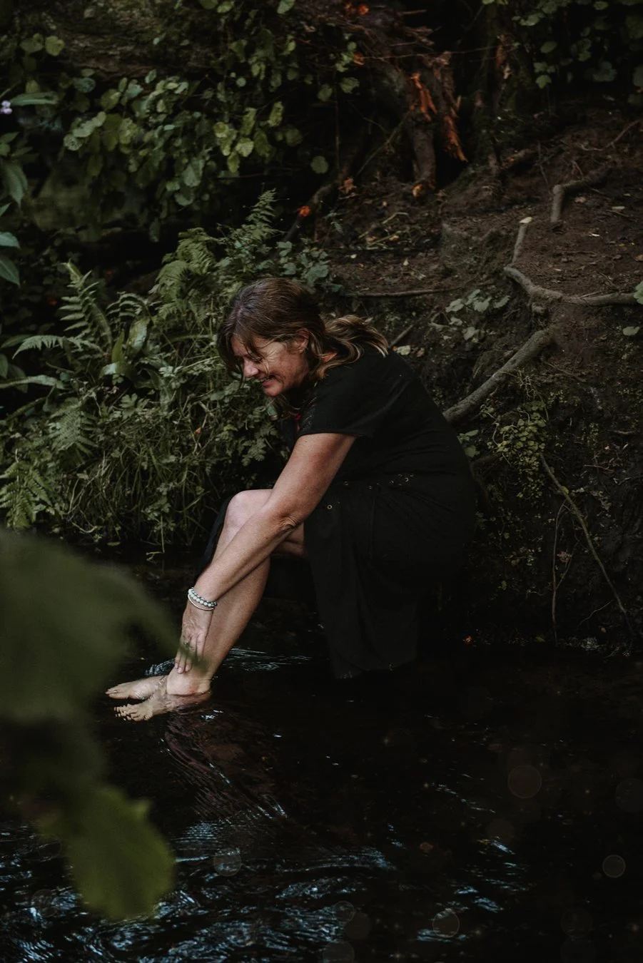 Paula from Tiny Rituals, she is leaning forward touching her leg while her feet are in the river water. She is surrounded by greenery.