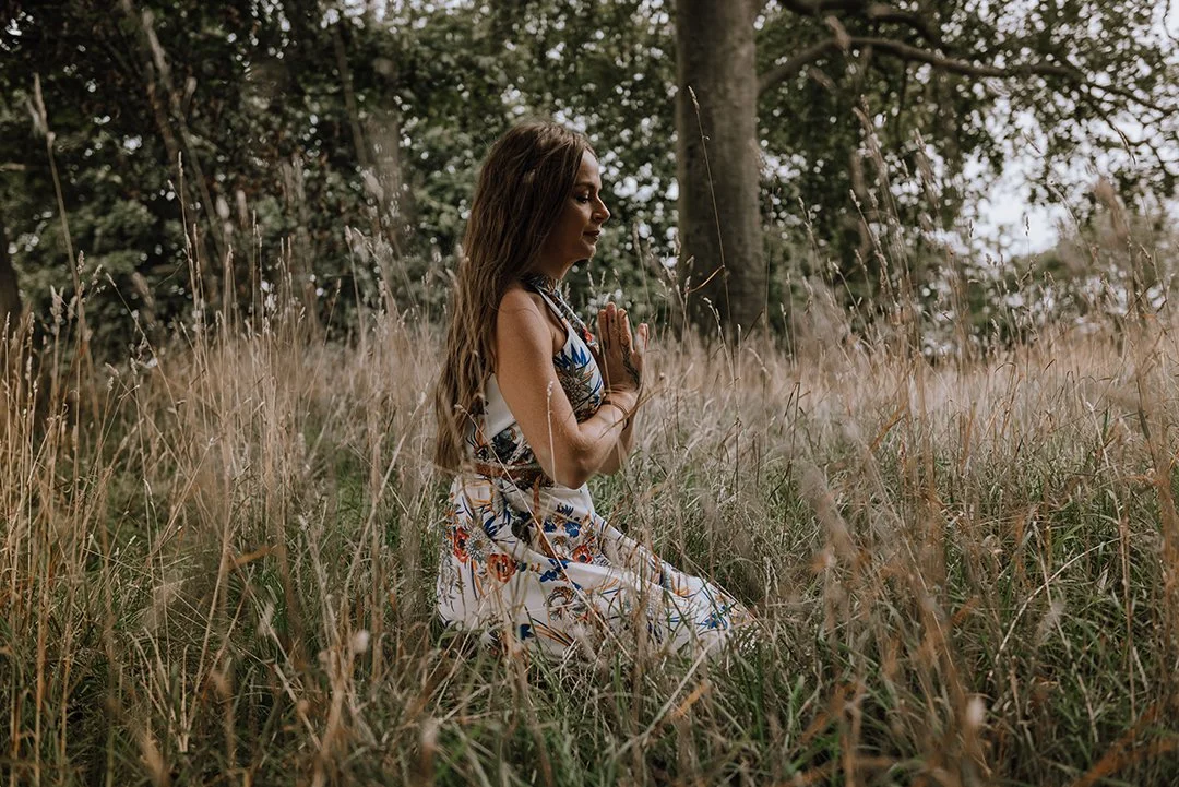 Holly Hollins, Higher Frequency coach. She is sitting amongst tall grasses, facing to the right side, her hands are in prayer position in front of her.