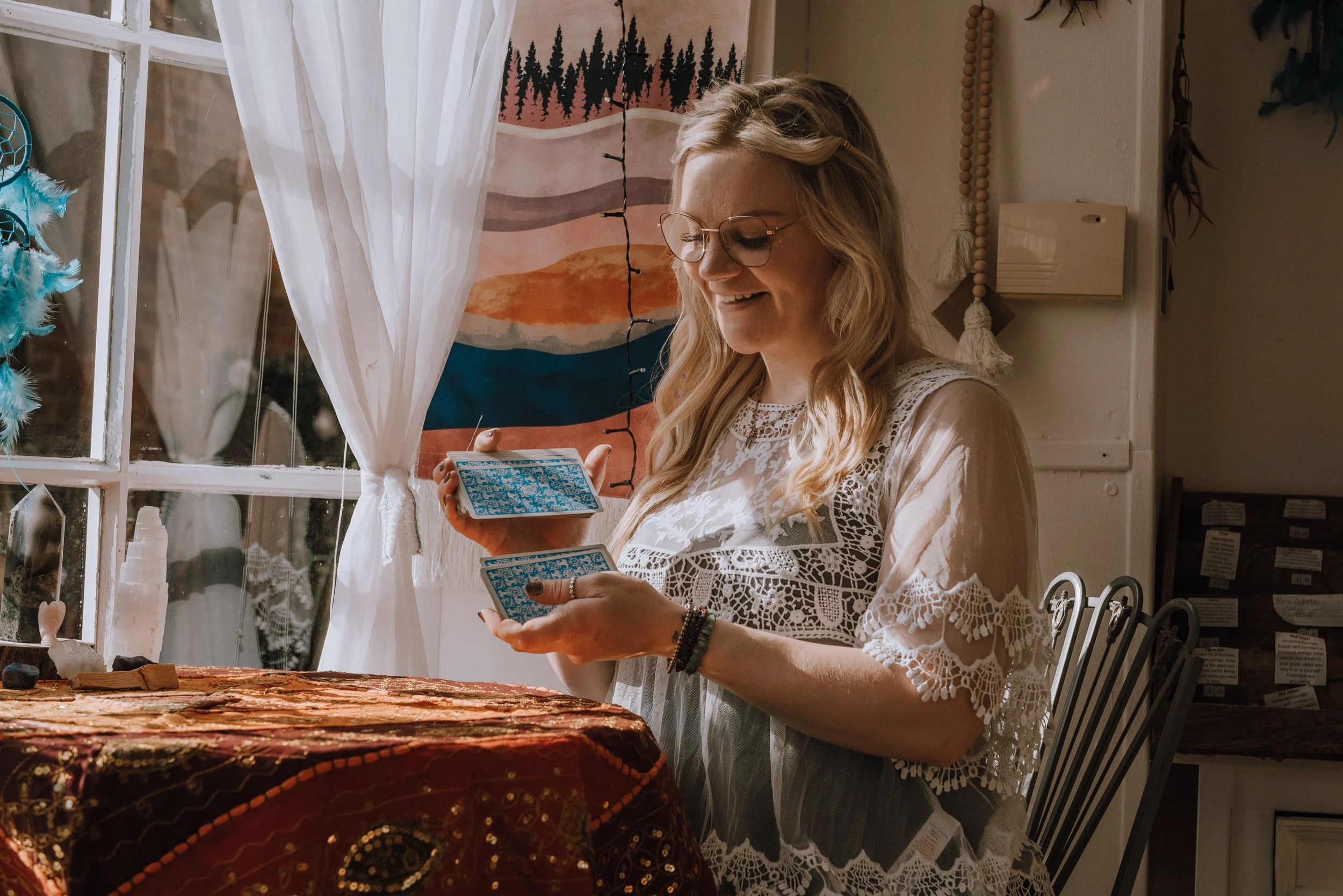 Camilla. Soul Reiki. She is shuffling Tarot cards while sitting at a table by a window. She looks happy. 