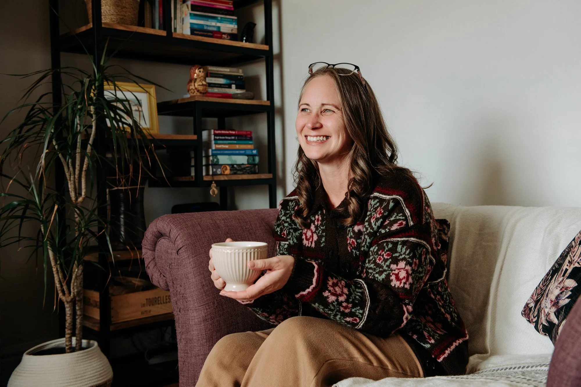 Jen, The Therapy Nook. She is sat on a comfortable sofa, holding mug. She is smiling to the side as if talking to a client.