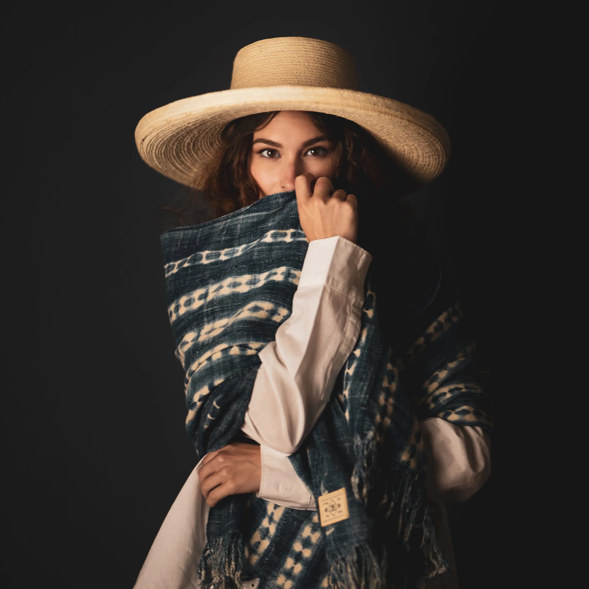 Woman wearing a large straw hat and patterned shawl, partially covering her face, against a dark background.