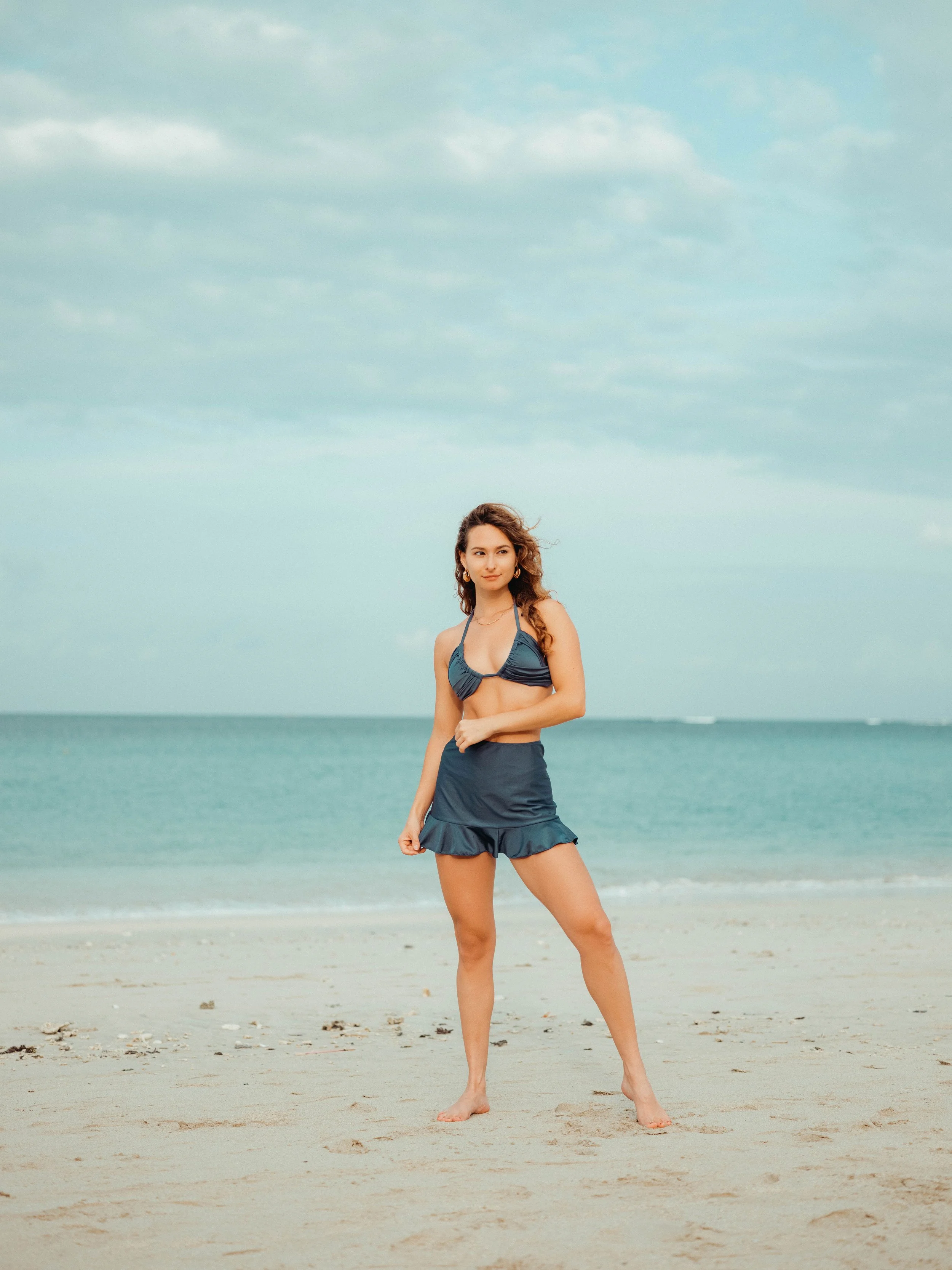 A woman standing on a sandy beach with the ocean and cloudy sky in the background, wearing a dark blue two-piece swimsuit.