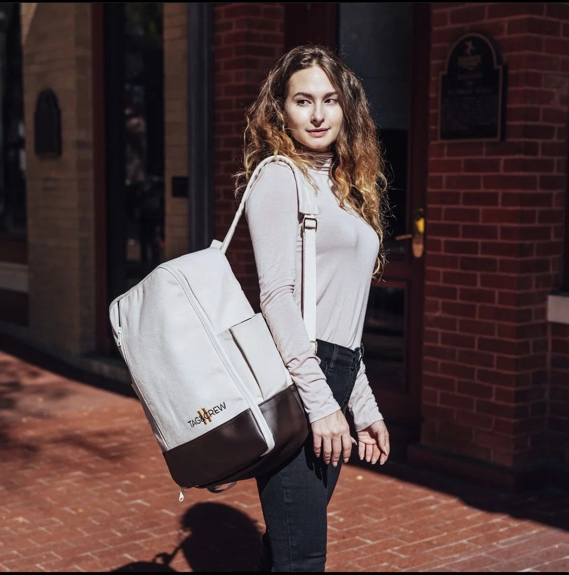 Young woman with curly hair carrying a white TAG Heuer crossbody bag standing on a brick sidewalk outside a red brick building at night.