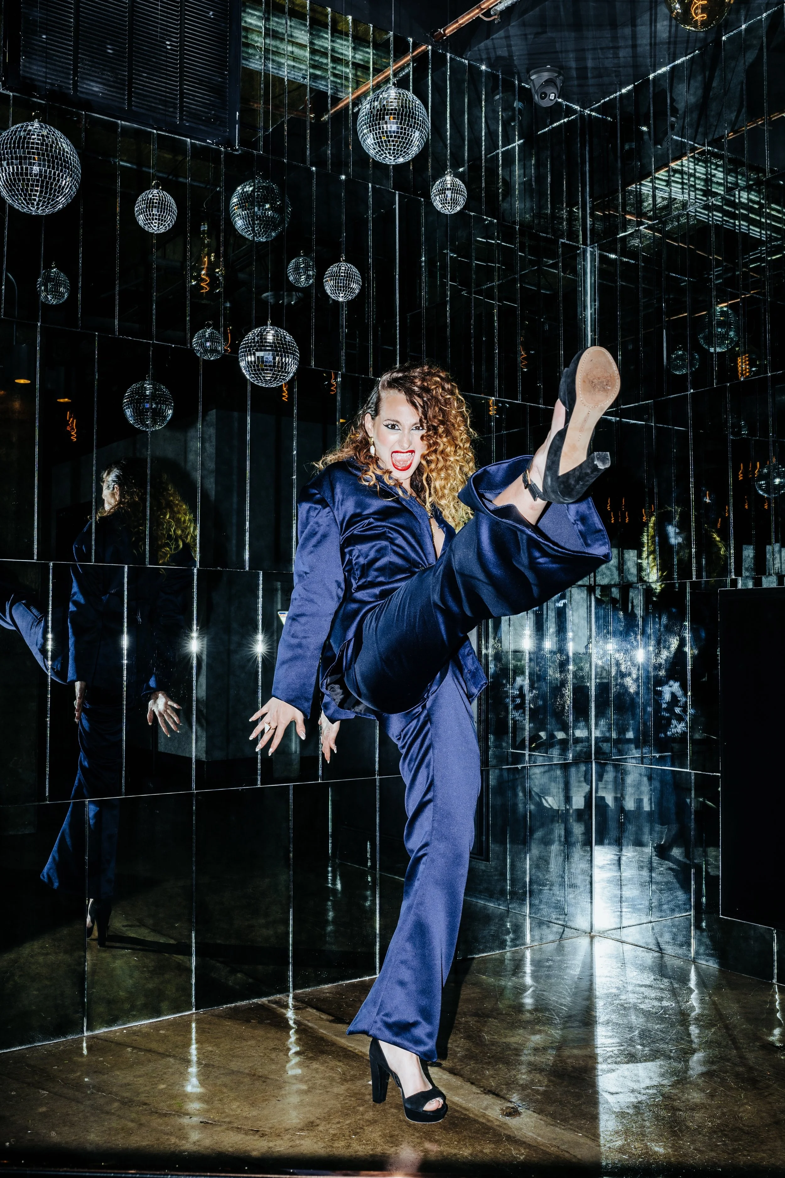 A woman in a dark blue suit and high heels performing a high kick in a mirrored room with disco balls hanging from the ceiling.