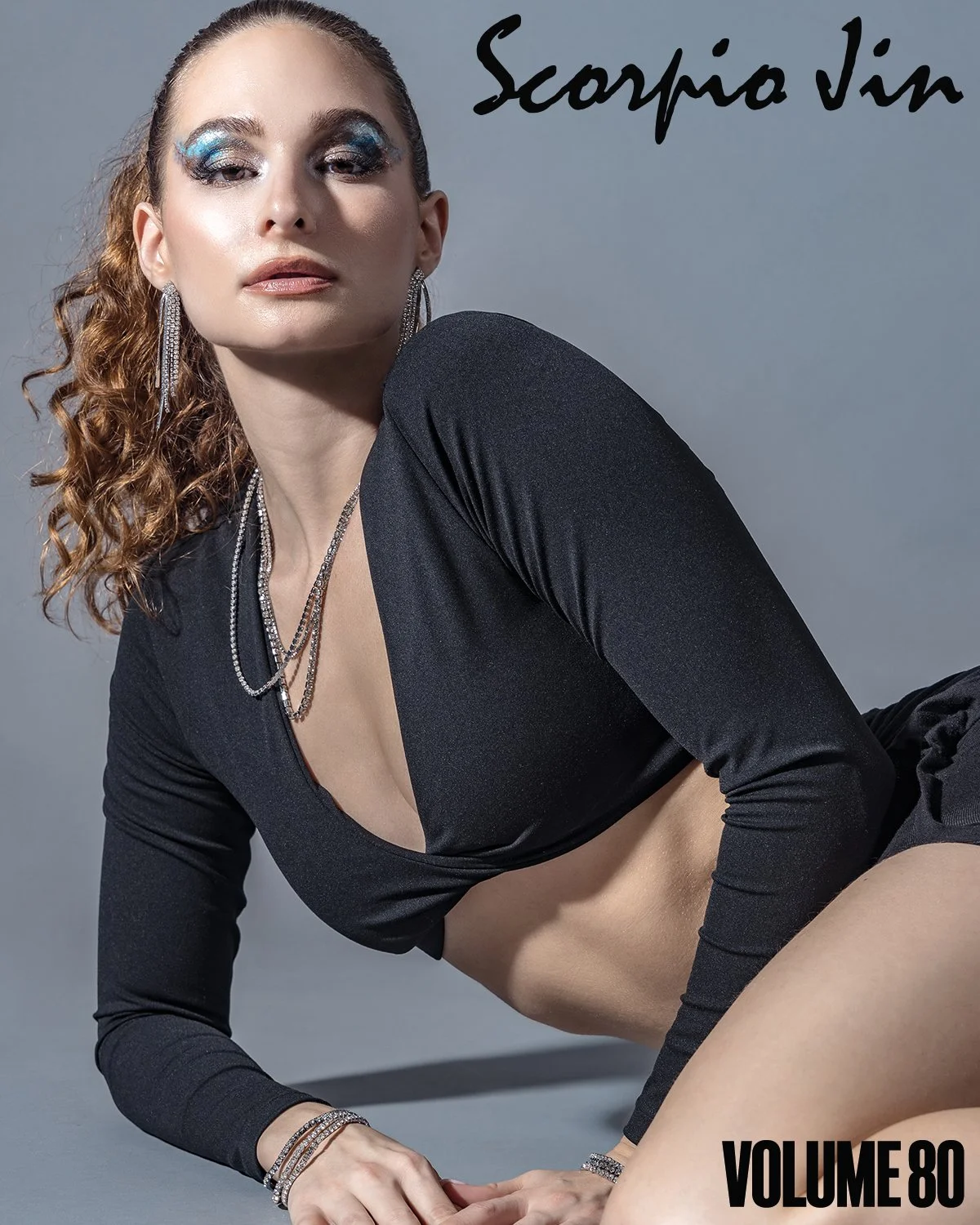 A woman with curly red hair wearing bold blue and silver eye makeup, jewelry, a black top with a plunging neckline, and black shorts, posing on the floor against a gray background with "Scorpio Sun" and "Volume 80" written on the image.