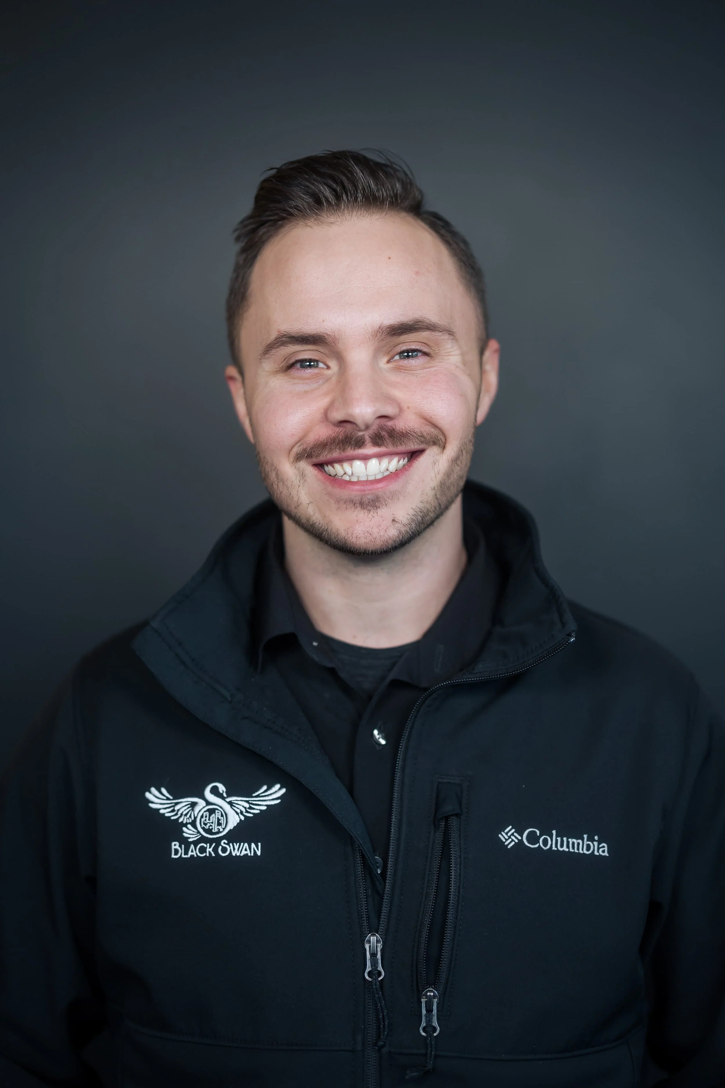 Professional headshot of smiling man in Black Swan Living Columbia jacket against a dark background