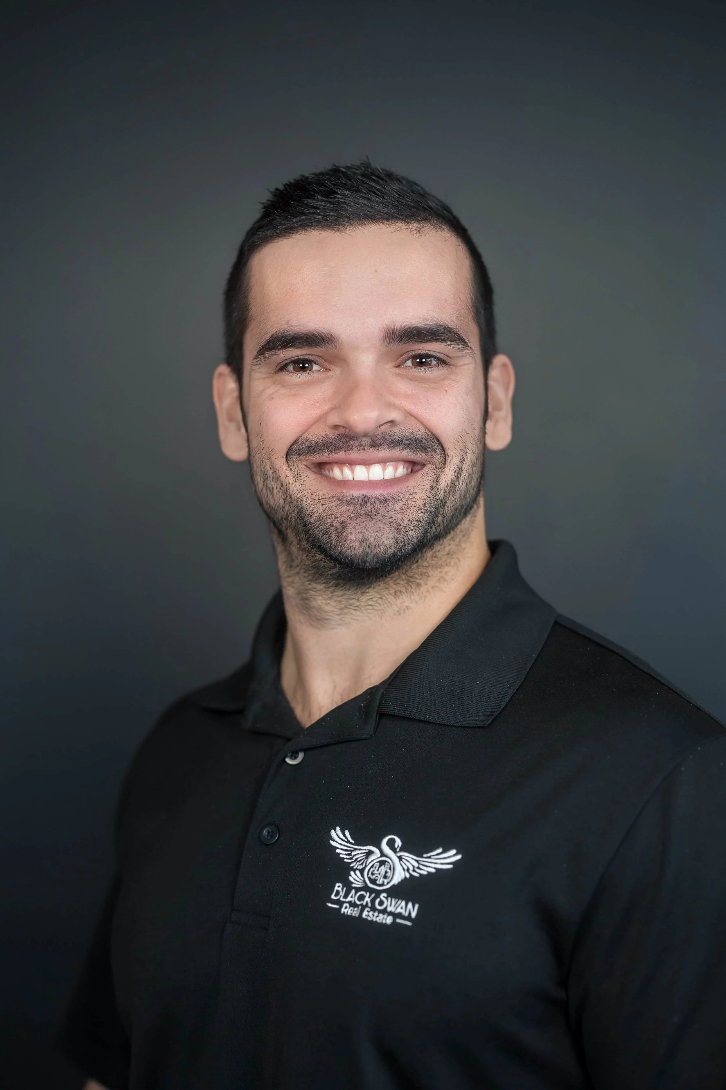 Professional headshot of smiling man in Black Swan Real Estate polo shirt on dark background