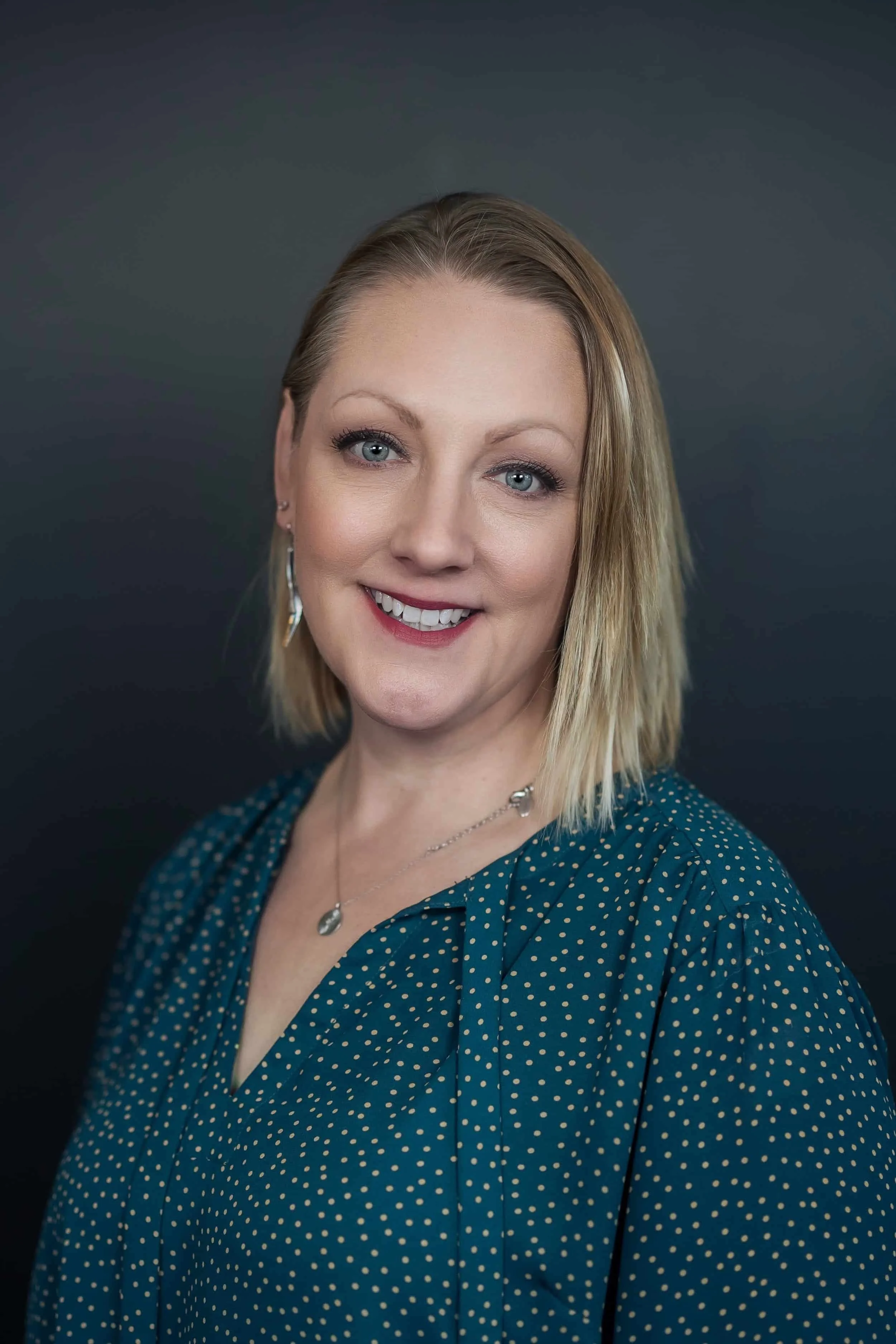 Professional headshot of smiling woman in teal polka dot blouse with necklace, posed against a dark background