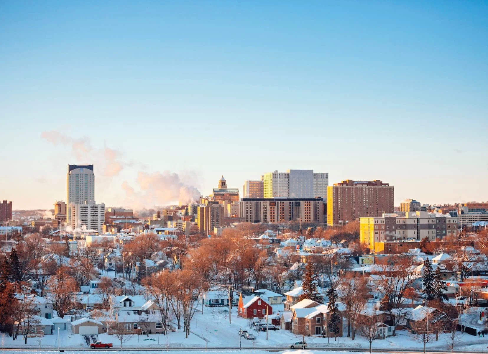 A city skyline in Rochester with tall buildings and houses blanketed in snow under a clear blue winter sky.