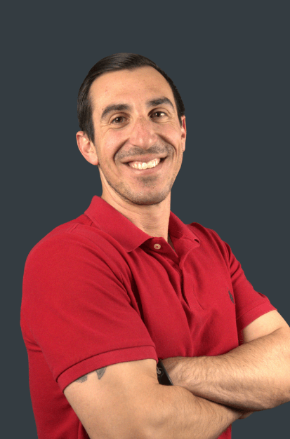 Professional headshot of smiling man in red polo shirt with arms crossed, posed against dark background