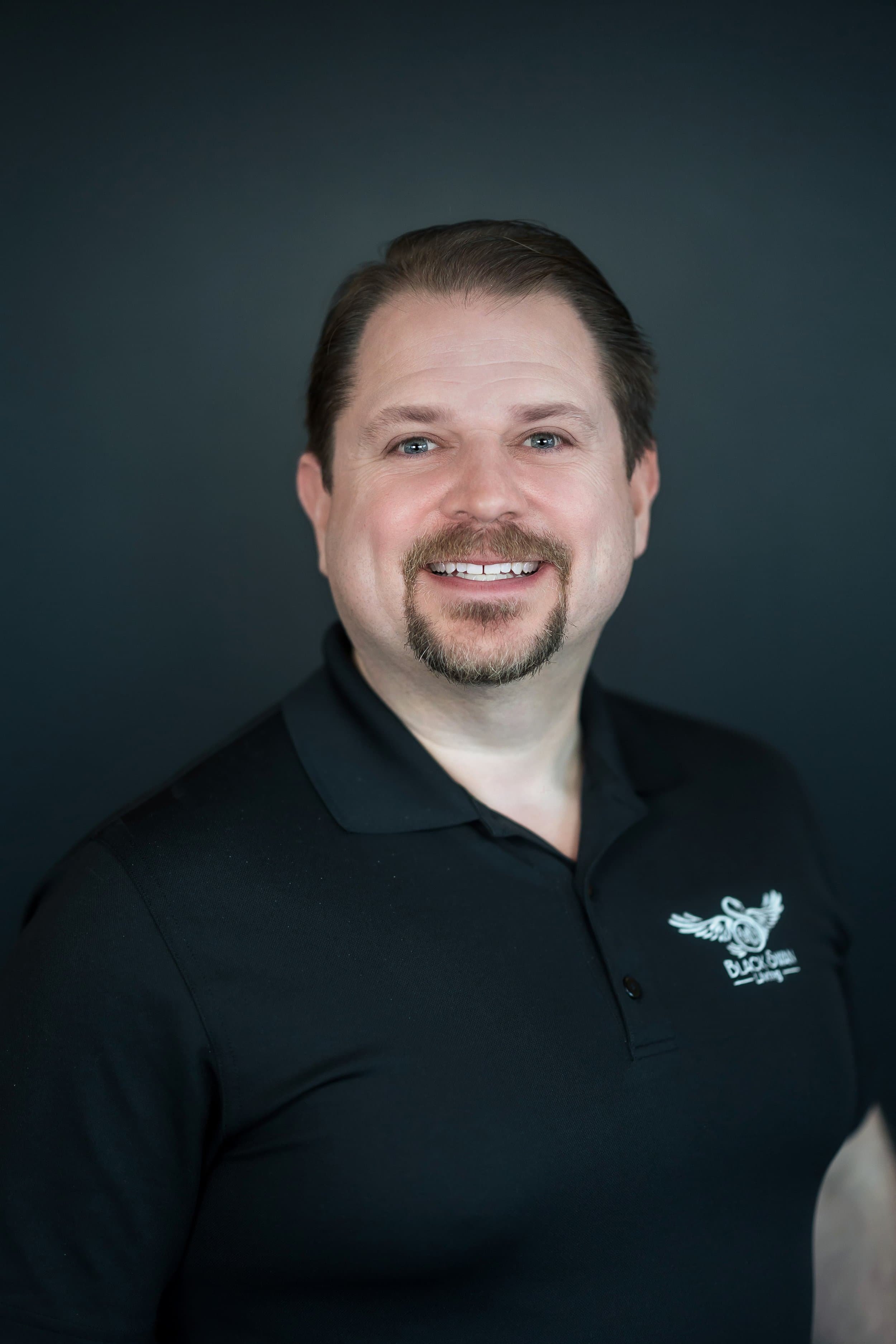 Professional headshot of smiling man in black Black Swan Living polo shirt on dark background