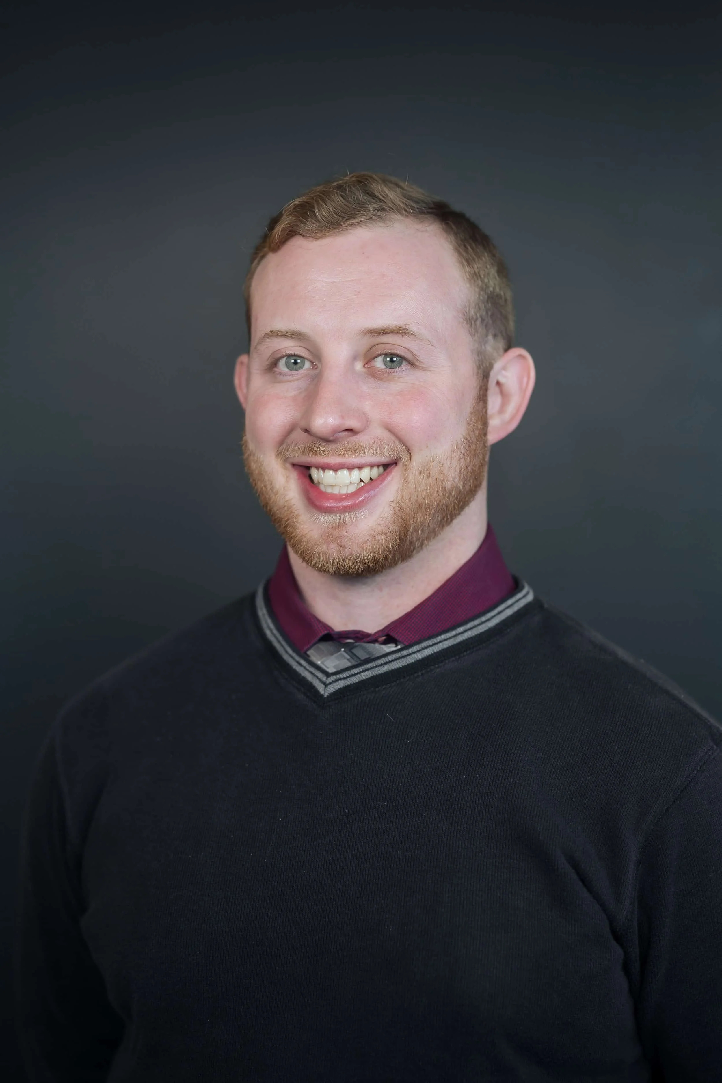 Professional headshot of smiling man in maroon shirt and black sweater with striped collar, posed against a dark background