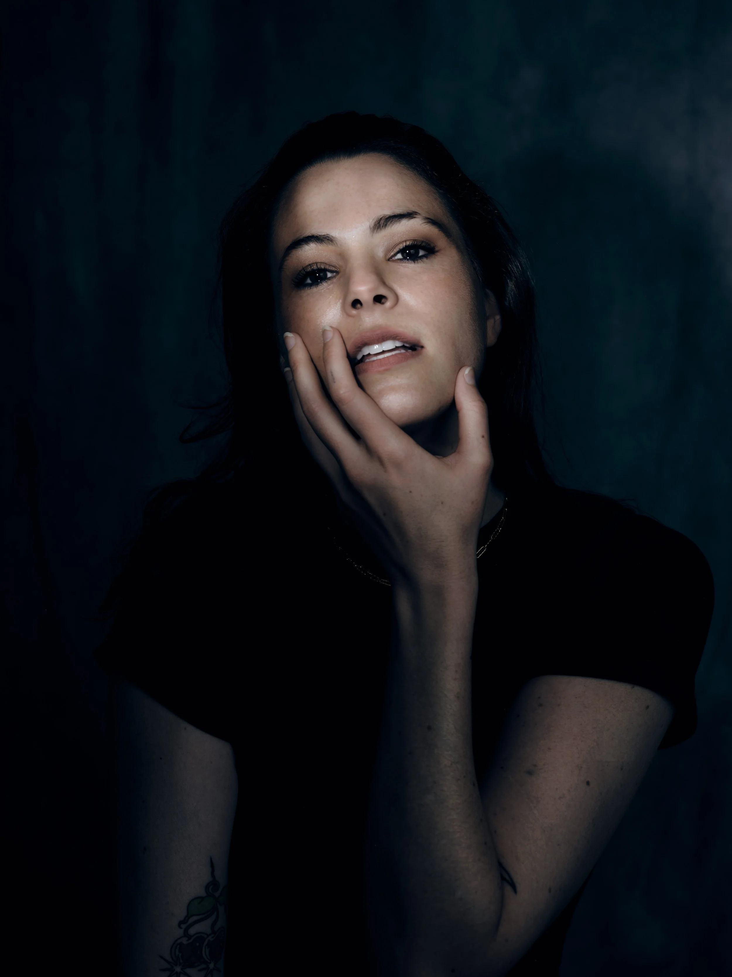 A woman with dark hair and fair skin holding her face with her hand against a dark backdrop.