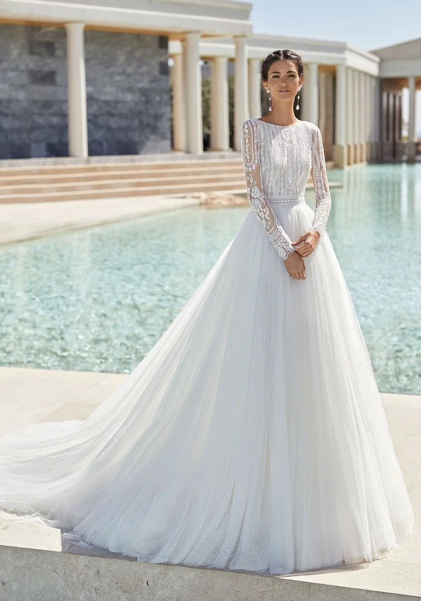 A woman wearing a white wedding dress with long lace sleeves, standing outdoors near a pool with classical-style columns and a modern building in the background.