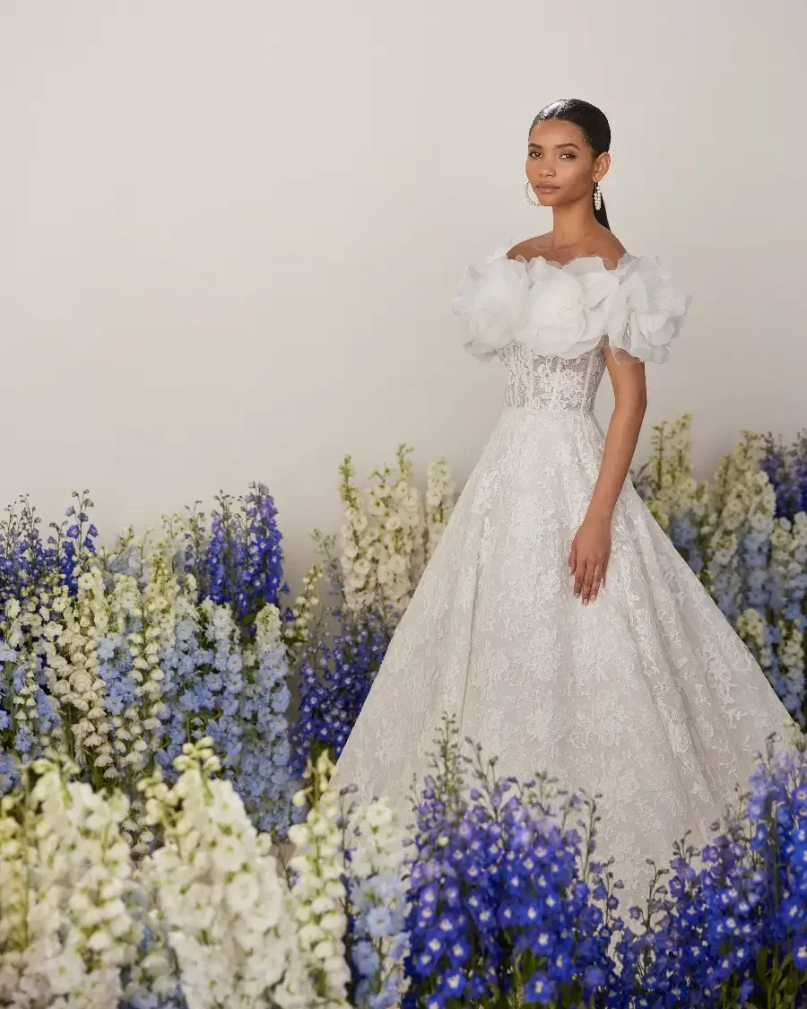 Beautiful bride standing in a hallway surrounded by groups of flowers in rebrode lace A-line  Ballgown with wrap at Ashley and Alexandrias Bridal Atelier Bloomfield Hills Michigan