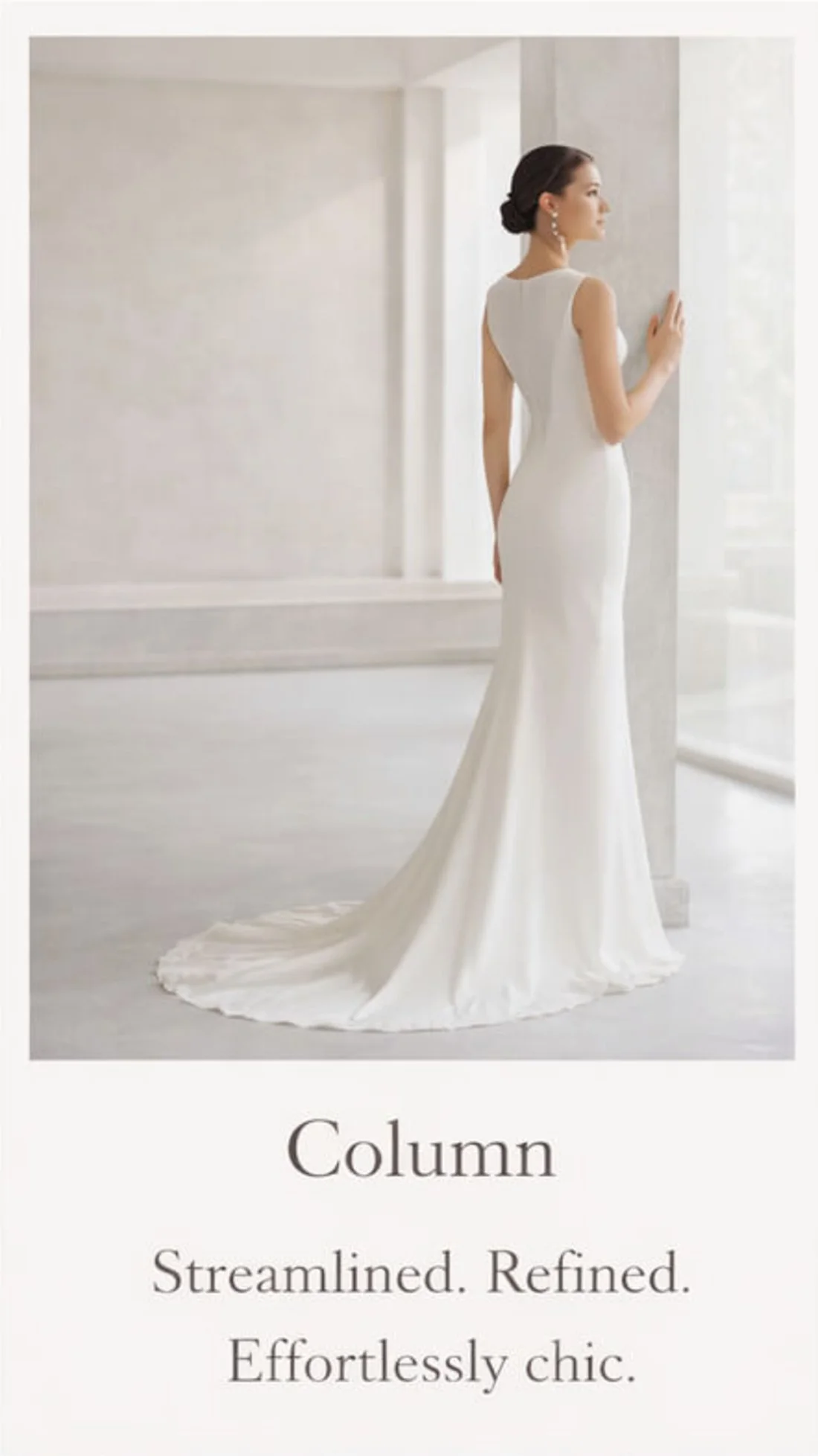 A woman in an elegant white wedding dress standing by a large window in a bright, minimalist room.