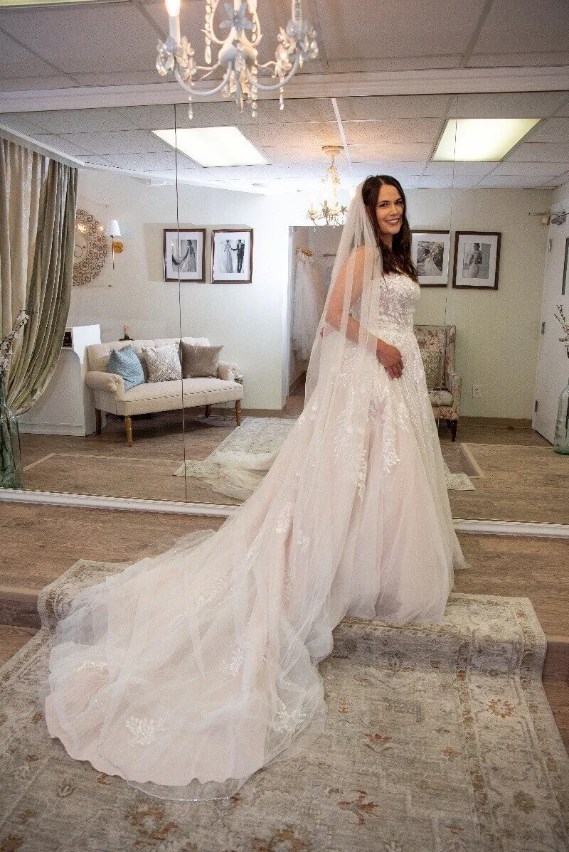 A woman in a wedding dress trying on her gown for a fitting, standing in a room with a large mirror, chandeliers, and framed black-and-white photographs on the wall.