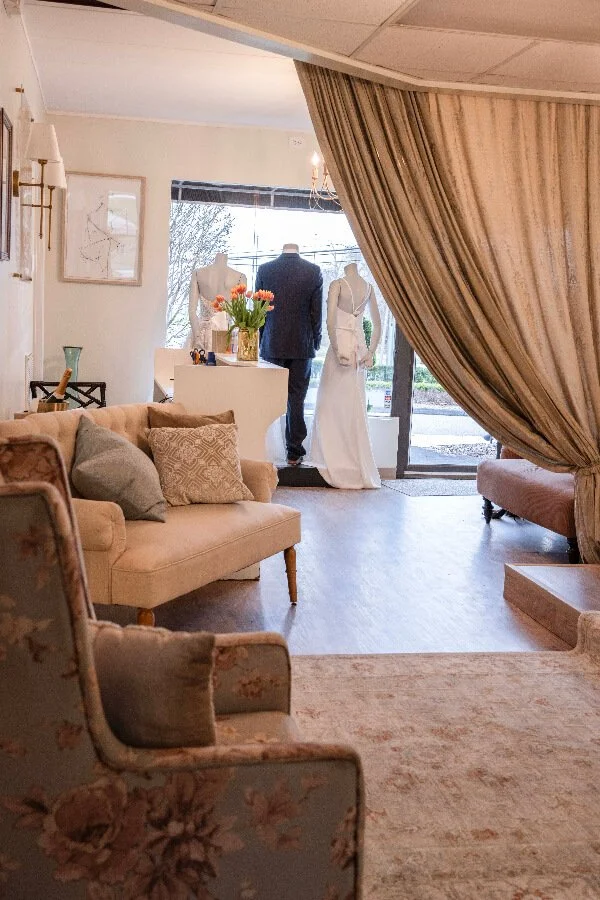 A cozy living room with a beige sofa and floral armchair, decorative pillows, a vase with flowers, and a curtained entryway showing mannequins dressed in wedding gowns inside a boutique.
