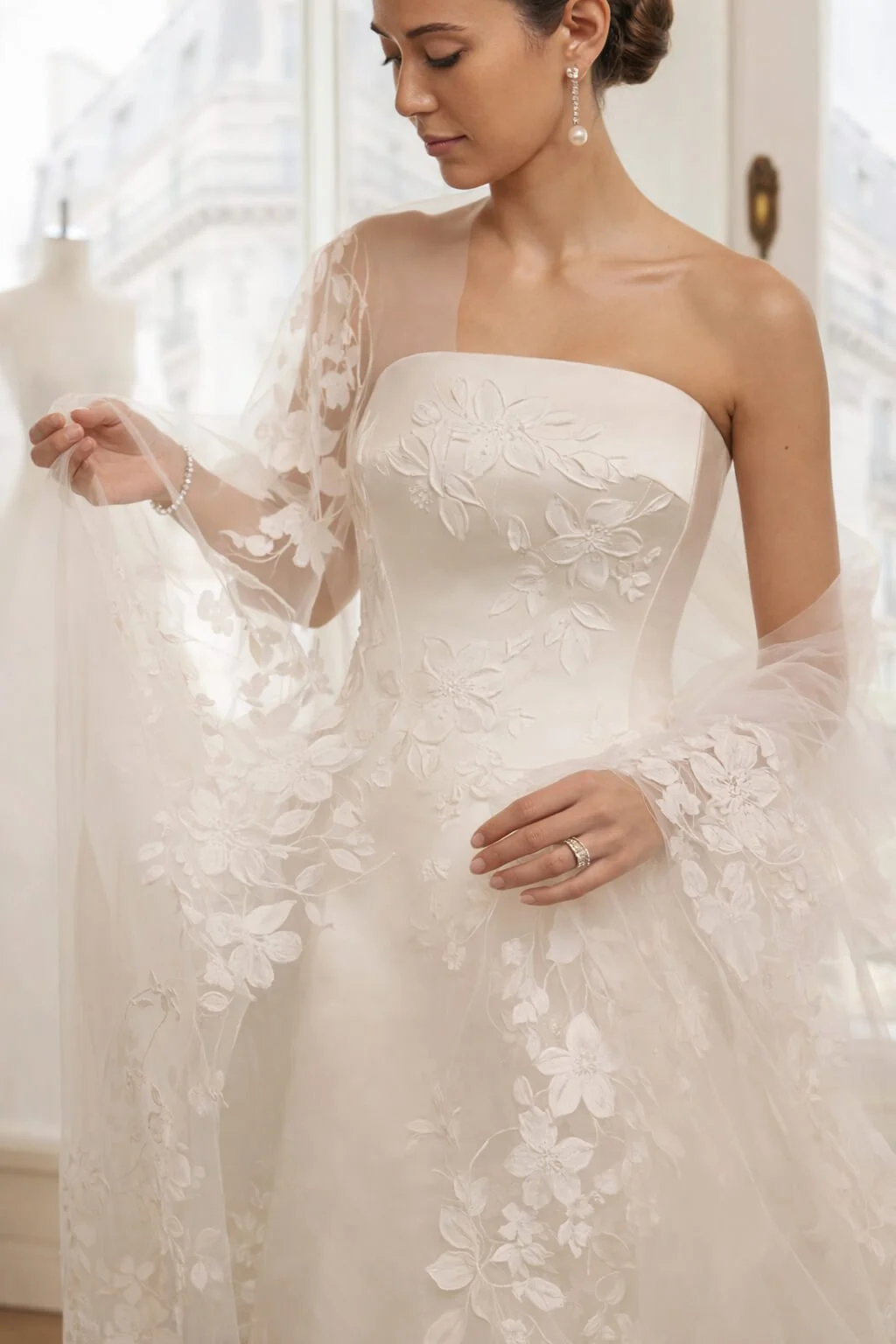 A woman wearing a white wedding dress with floral embroidery and sheer lace sleeves, standing indoors with a cityscape visible through the window behind her.