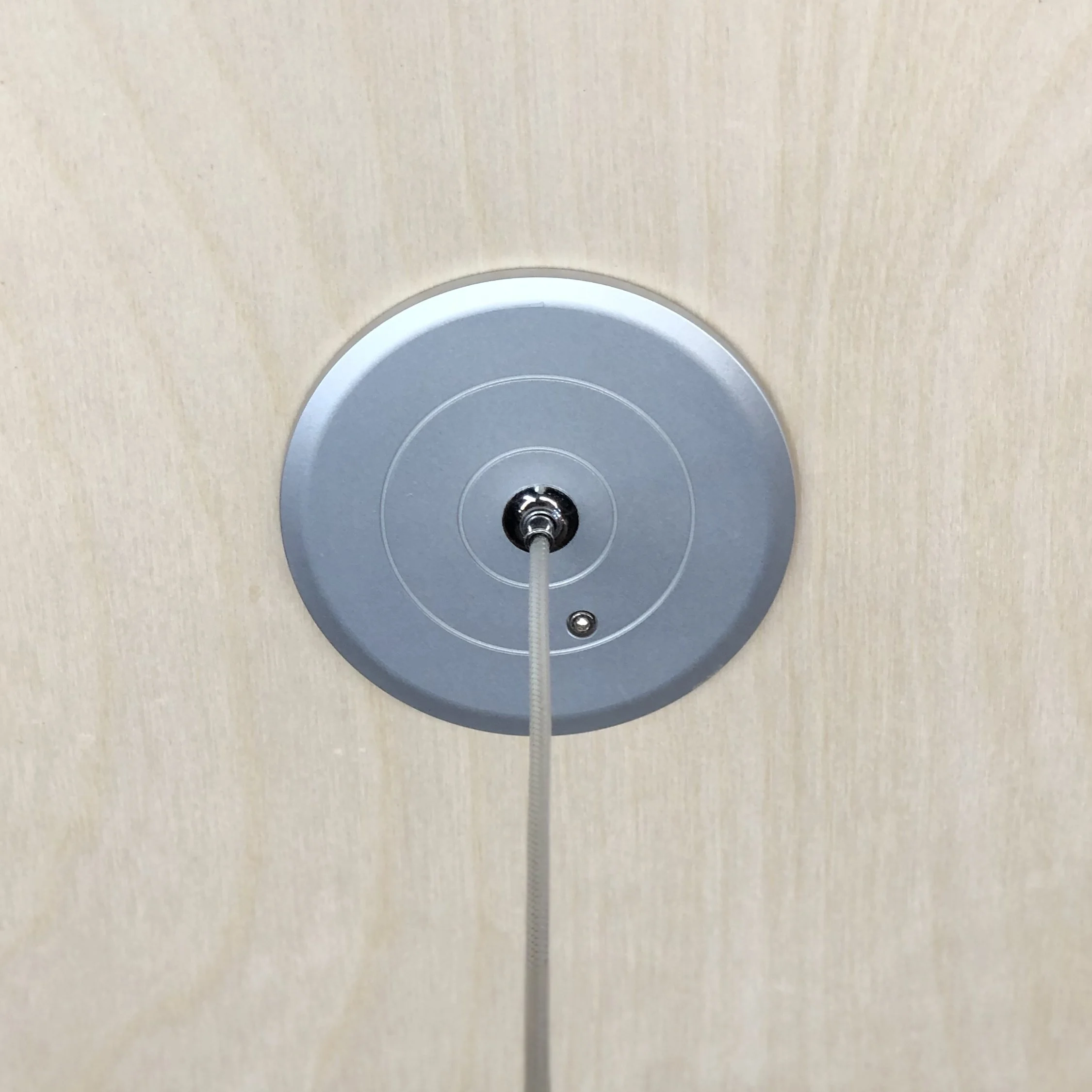 Close-up view of a circular metallic fixture, possibly a ceiling light or smoke detector, mounted on a beige textured ceiling.