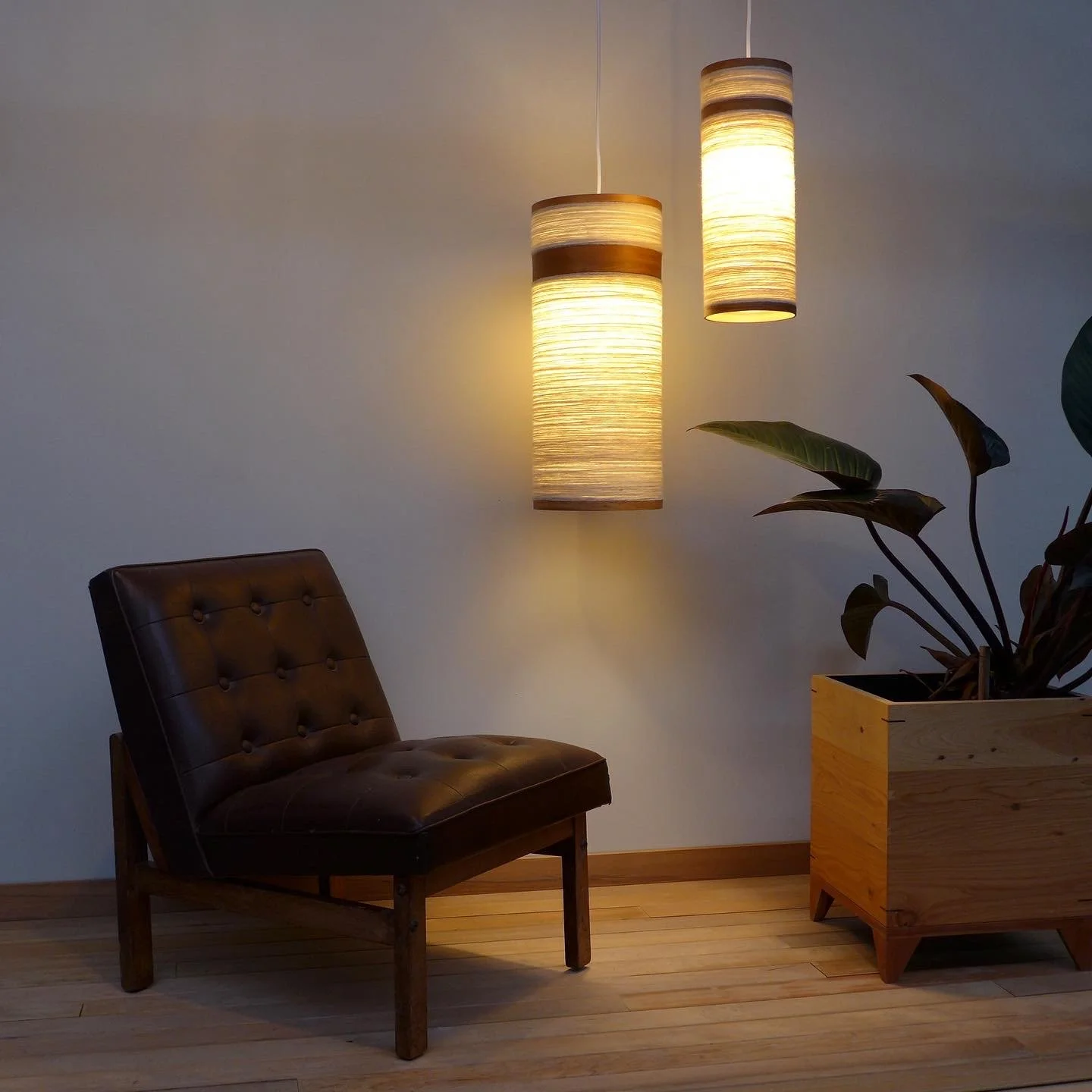 A cozy corner with a leather chair, two hanging pendant lights, a potted plant in a wooden box, and a wooden floor.