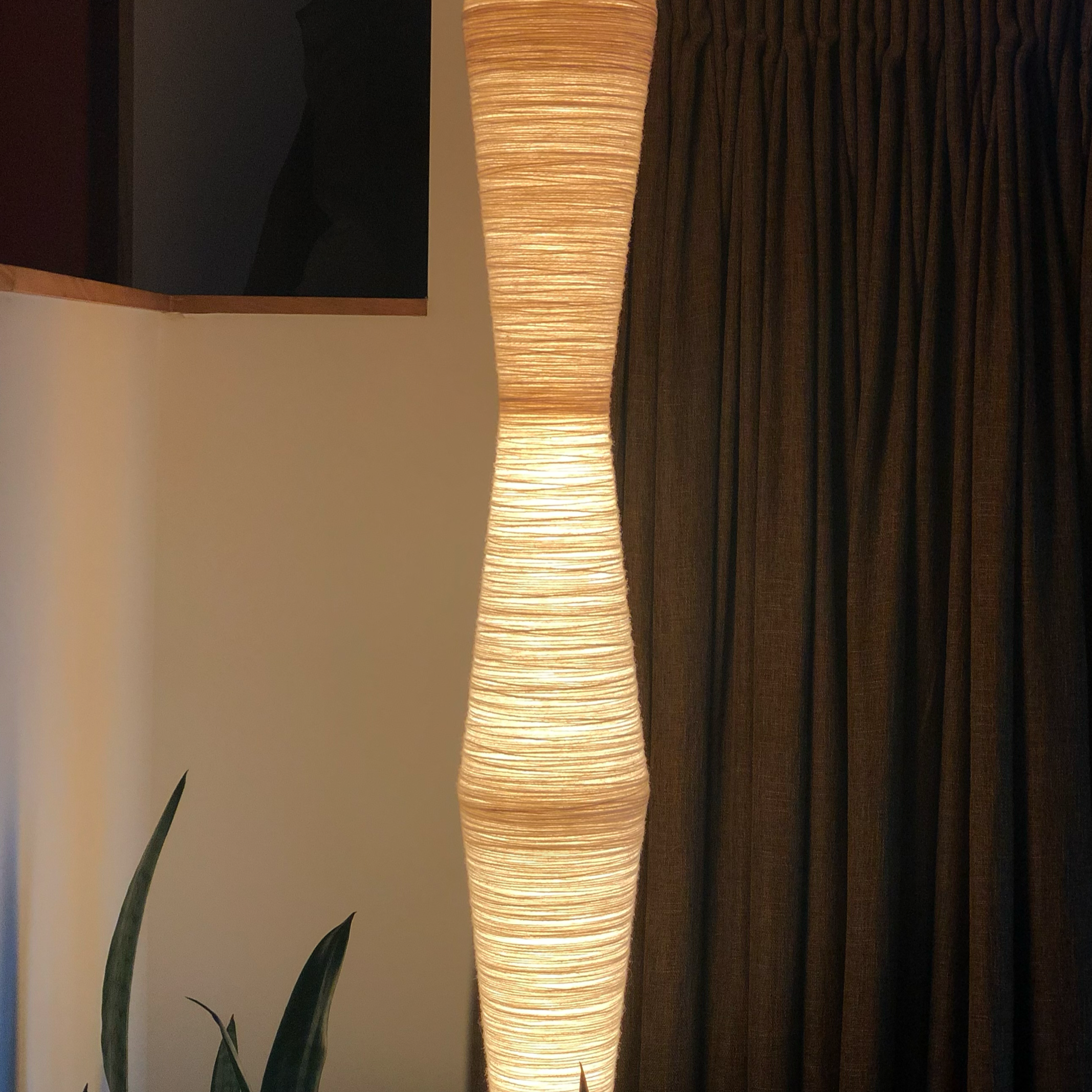 A tall, cylindrical light fixture with a woollen texture, emitting a warm glow, positioned next to a dark textured surface and a plant with long, pointed leaves.