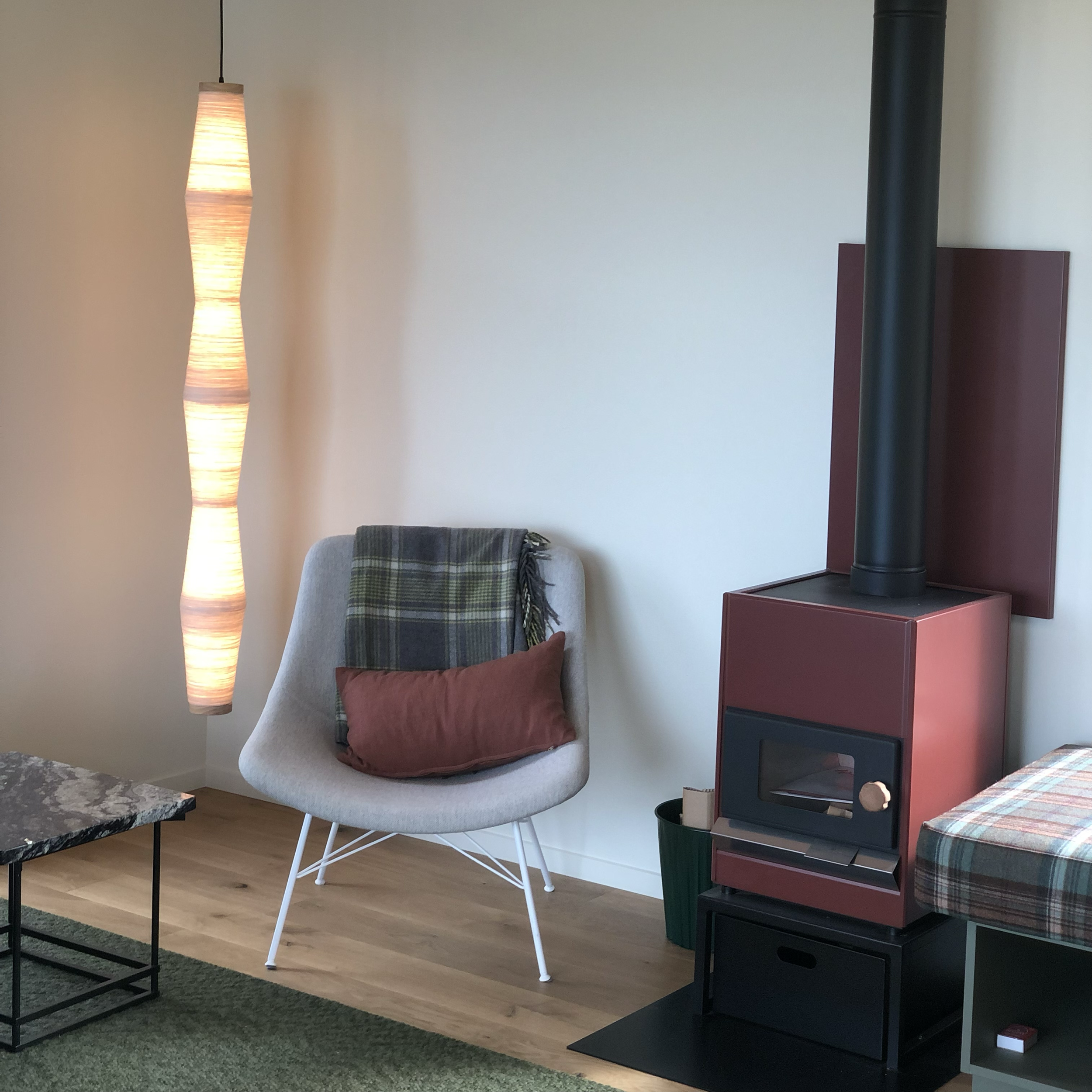 A cozy interior room with a modern hanging lamp, a beige armchair with white legs and a plaid throw, and a dark-colored wood stove on a red base unit. A small side table with books and a cubed decorative piece is seen in the foreground. The floor is 