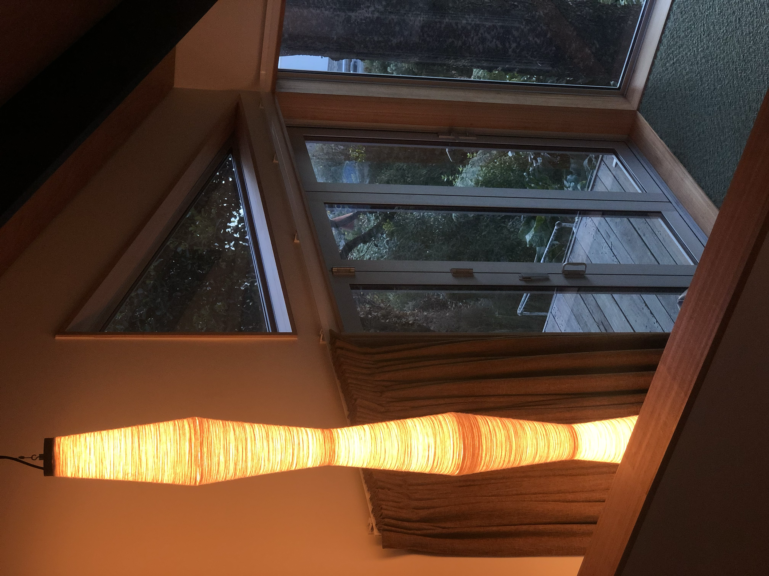 Interior view of a room with a tall decorative glowing pendant light, a large glass sliding doors leading to an outside deck with trees visible outside.