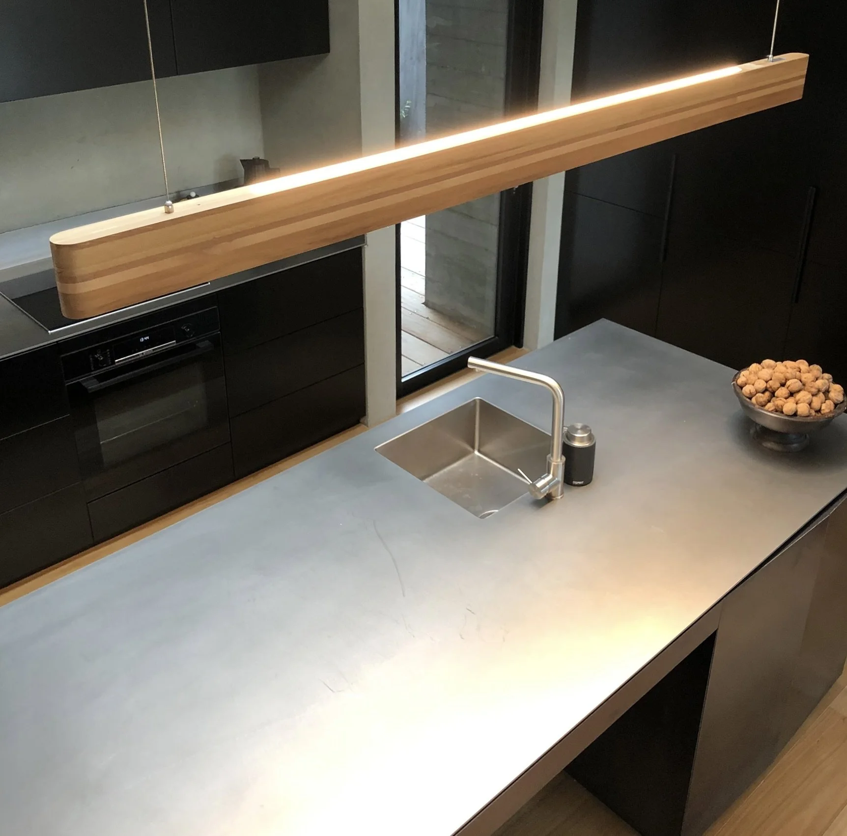 Modern kitchen with a marble countertop, a stainless steel sink with a tap, a bowl of walnuts, black cabinets, and a wooden ceiling light fixture.