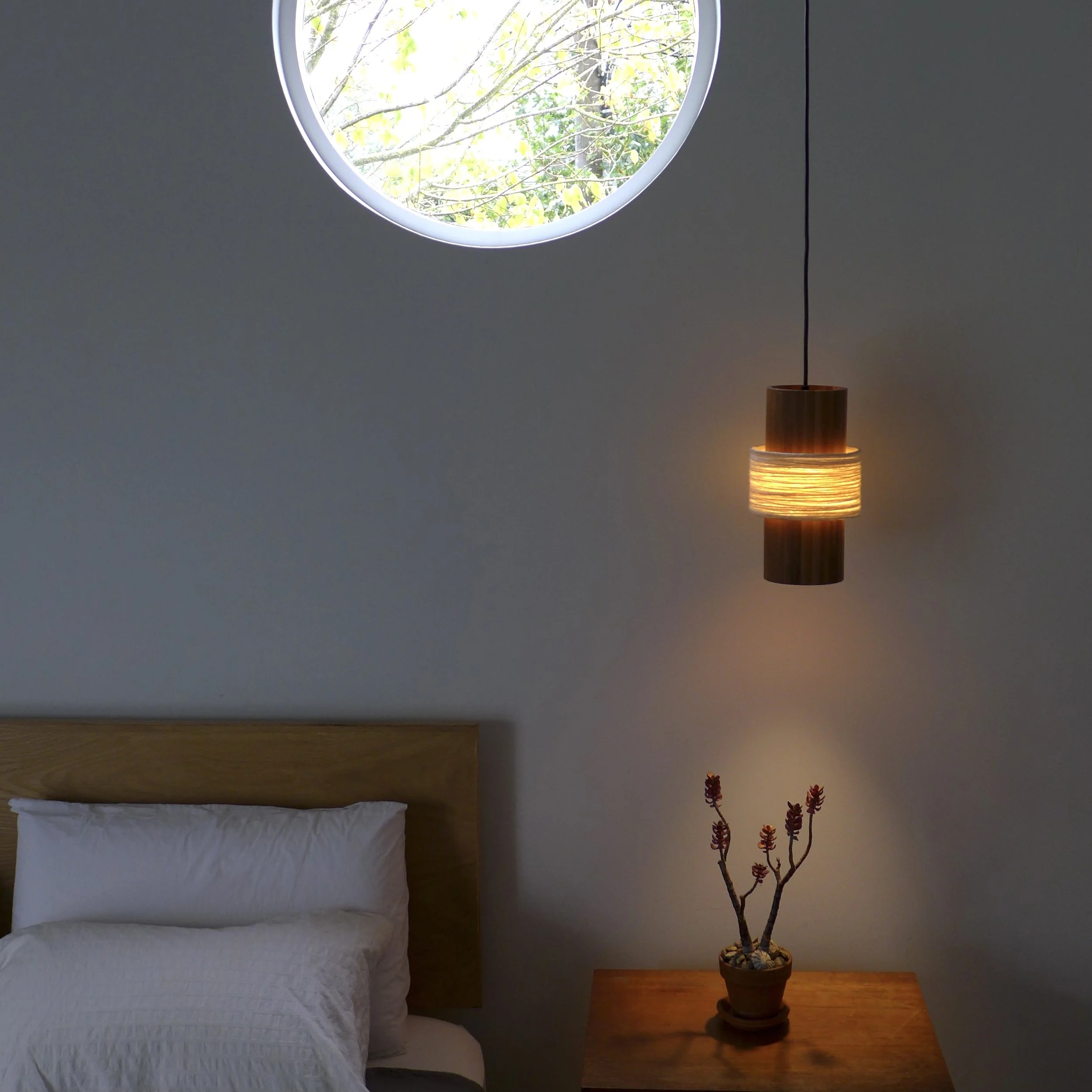 A bedroom scene with a bed dressed in white bedding in the lower left corner, a dark wooden nightstand on the right with a potted plant featuring purple flowers, and a window above the bed showing tree branches and yellow-green leaves outside. A mode