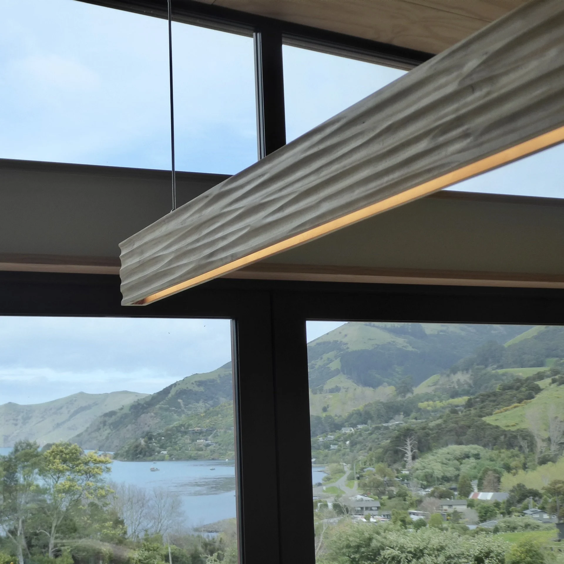Scenic view of a lake surrounded by green hills and trees, seen through large glass windows of a modern building.