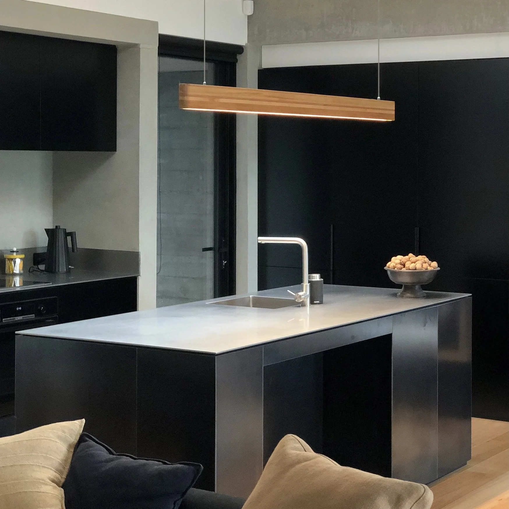 Modern kitchen with black cabinets, a white island with a sink, a bowl of nuts on the counter, and a wooden hanging light fixture. In the foreground, person's legs in beige pants and a dark sweater are visible, sitting on a couch.