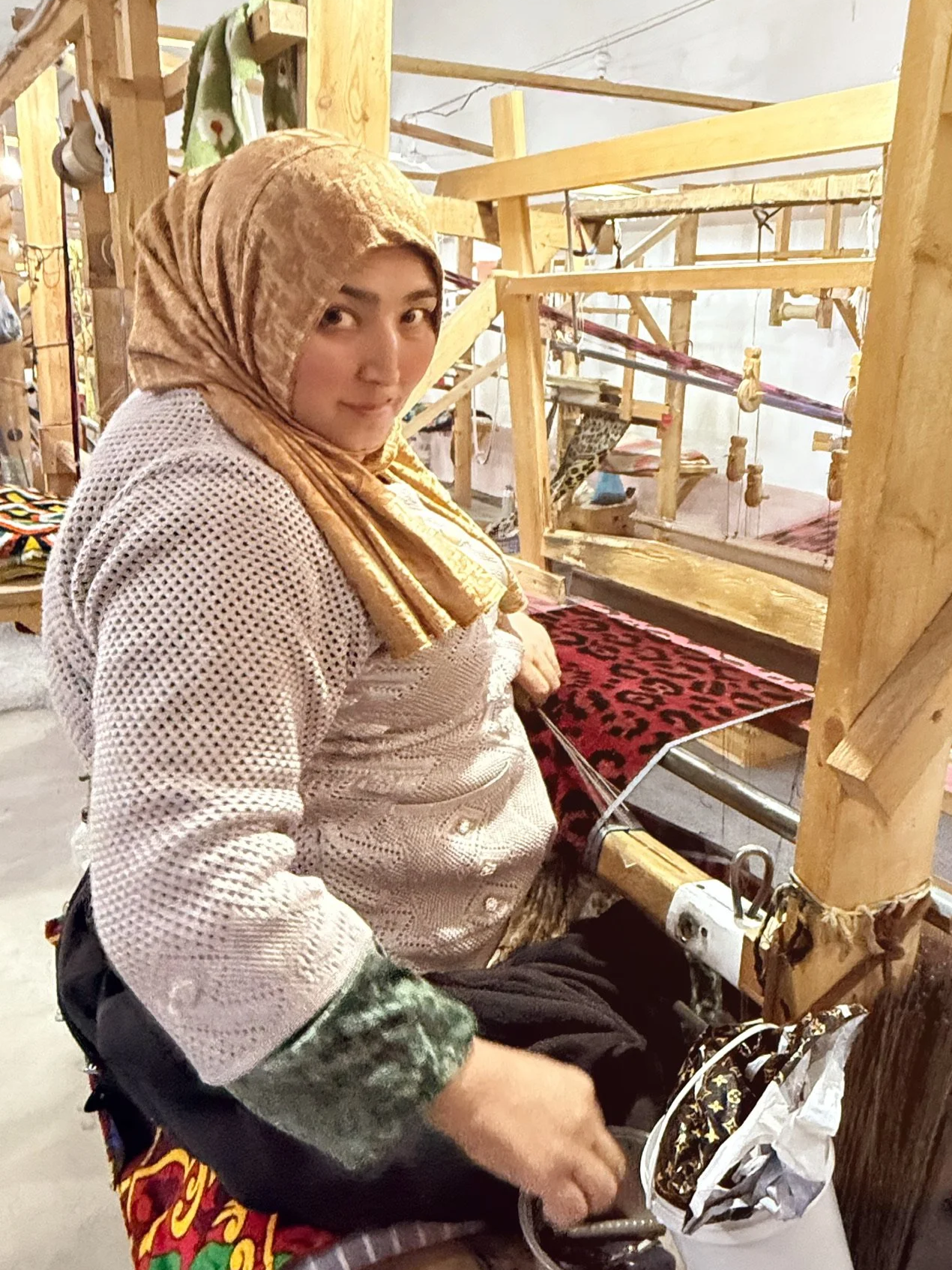 A woman in a gold hijab and a white crochet top sitting at a loom, working on a red and black patterned textile in a workshop.