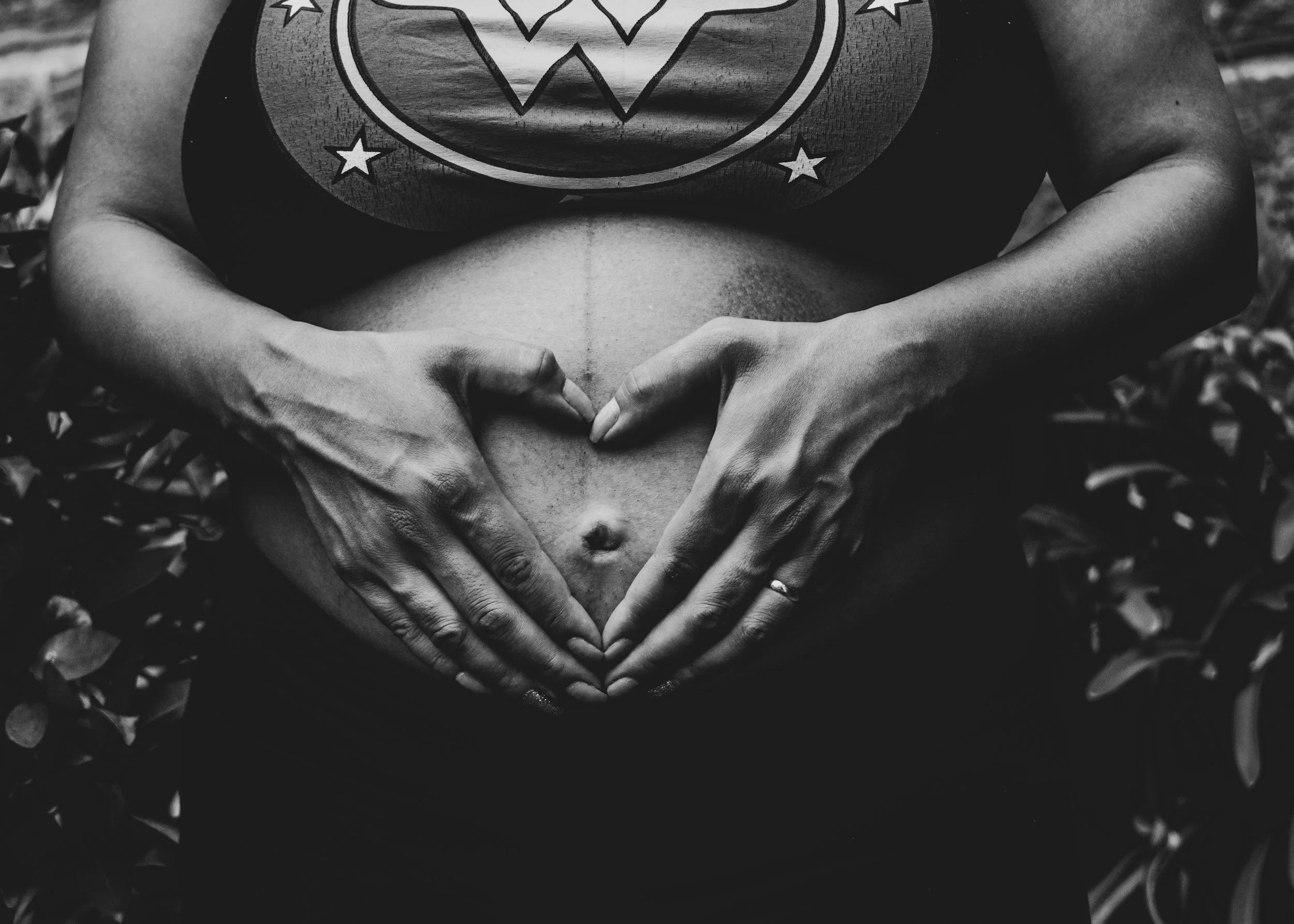 Black and white photograph of a pregnant person forming a heart shape with their hands on their abdomen, wearing a shirt with a Wonder Woman logo.